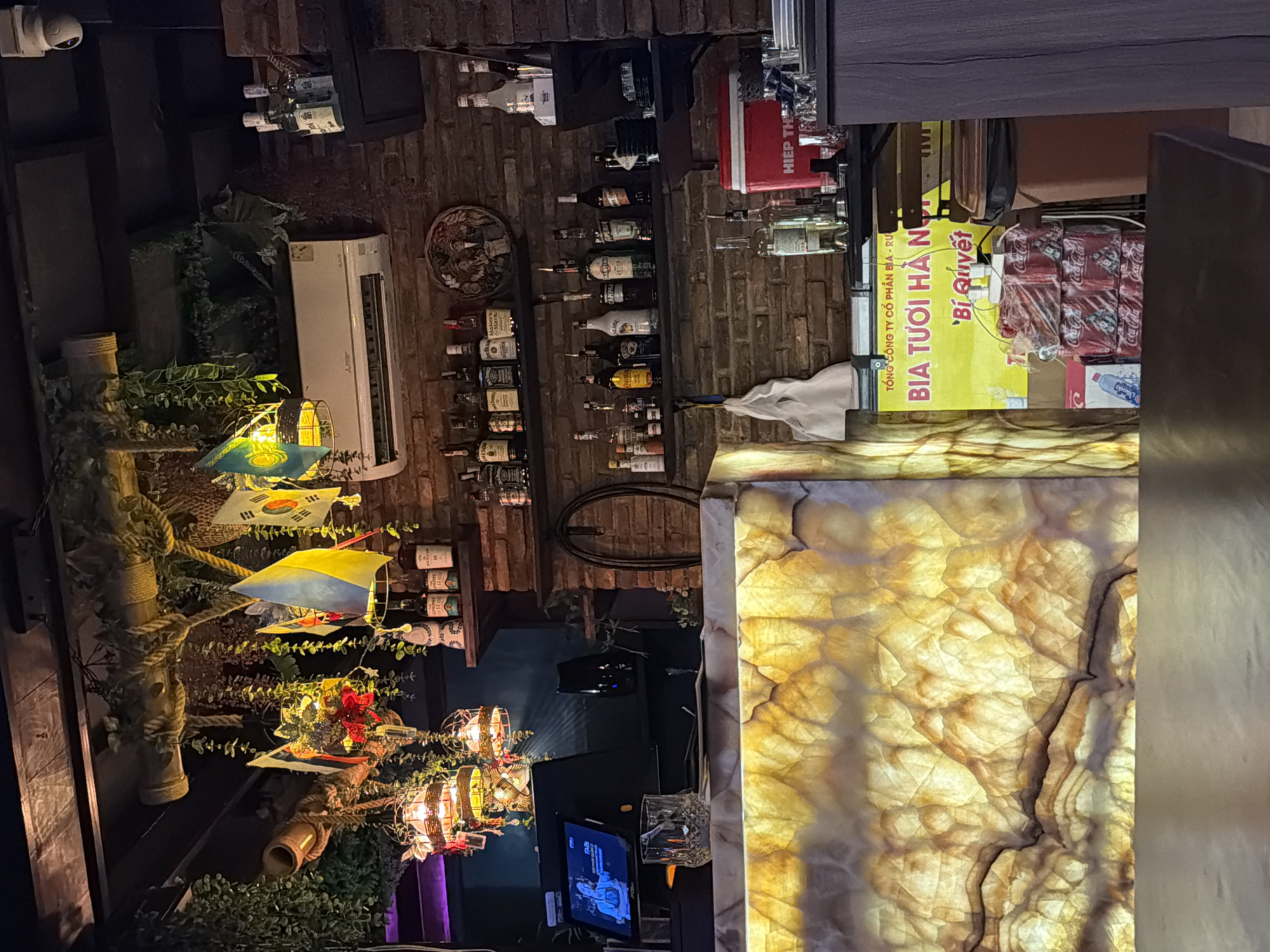 The interior of a bar in Nha Trang Ward, Vietnam, is captured, with a brick wall displaying a collection of liquor bottles. Hanging decoratively are flags of various nations. A modern air conditioning unit is mounted on the wall. A counter with a distinctive yellow marble pattern is visible. A television is broadcasting an image of people, adding an element of activity. The lighting is warm, contributing to the ambience.