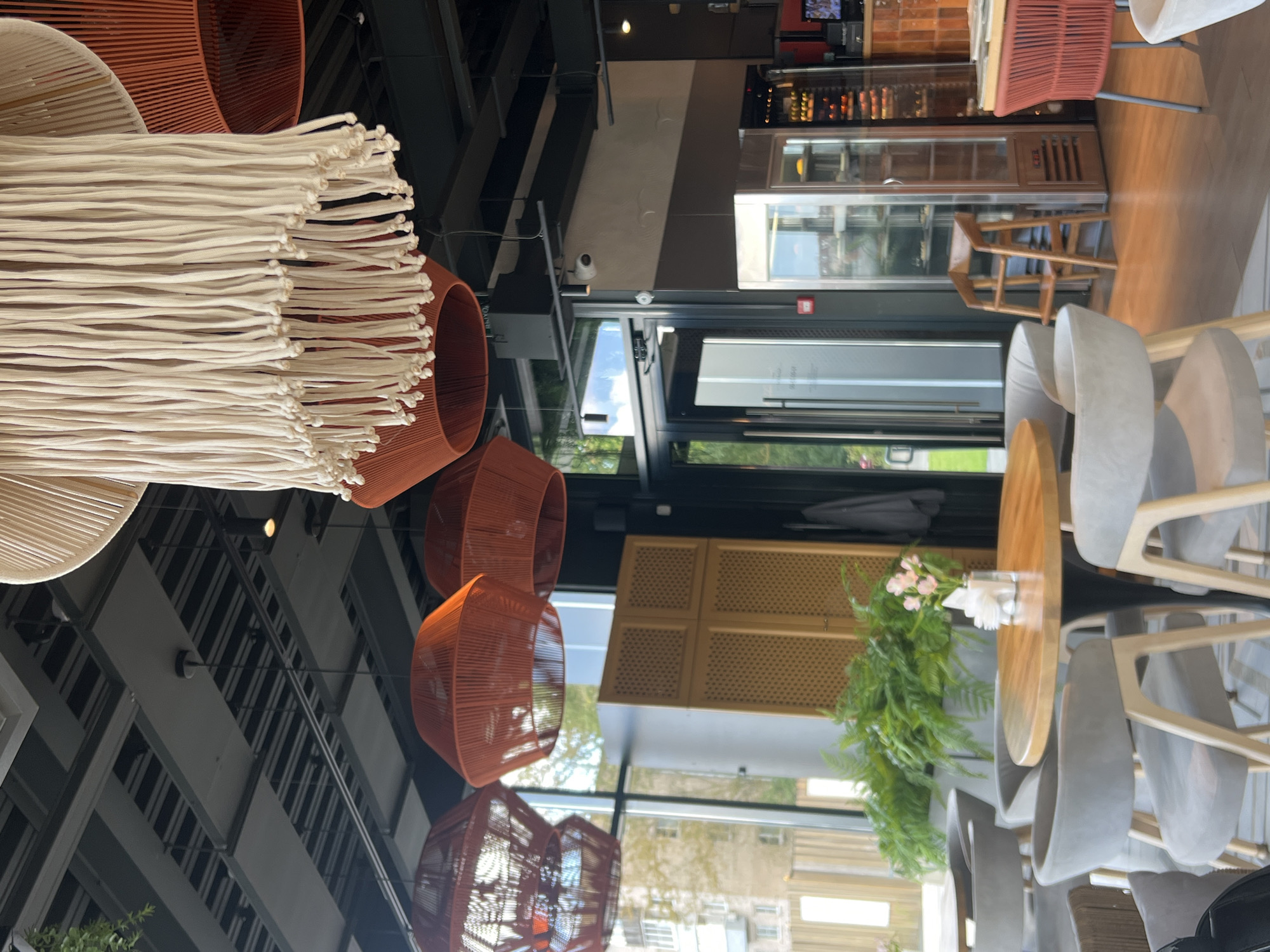 The image depicts the interior of a modern restaurant or cafe, likely in Veliky Novgorod, Russia, given the context. The scene is brightly lit, with a focus on the ceiling decorations and dining area. Hanging from the ceiling are large, distinctive lampshades. One prominent lampshade is made of white, braided ropes, creating a fringed effect. Below this, several large, conical lampshades in a vibrant terracotta or burnt orange color are visible.
The dining area features several round and square tables with contemporary chairs. The chairs are upholstered in a light gray fabric with wooden legs. One round table in the foreground has a light wooden top and a dark base, set with a small vase of pink flowers and napkins. Large windows line one side of the establishment, offering a view of trees and buildings outside, suggesting an urban or suburban setting.
Further into the restaurant, a glass-fronted display refrigerator, possibly for food or beverages, is visible. Next to it, a wooden high chair for a child is positioned. The flooring is polished wood. The overall aesthetic suggests a stylish and comfortable dining environment. There are no people clearly visible in the image, making it a static portrayal of the venue.