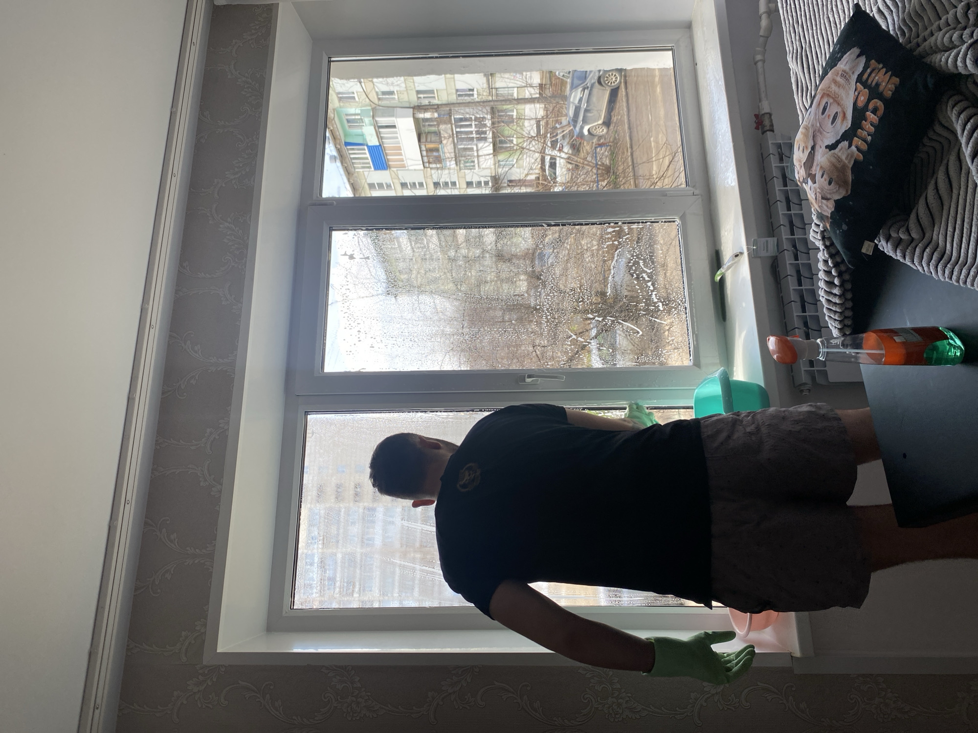 A man is cleaning a window in an apartment in Chaikovsky, Russia. He is wearing a black t-shirt, khaki shorts, and green gloves. The window is divided into three panes, and the middle pane is covered with soap. Outside the window, apartment buildings and trees can be seen. A cushion with text reading "TIME TO CHILL" sits behind the man. A radiator is also visible, along with a spray bottle and a bucket.