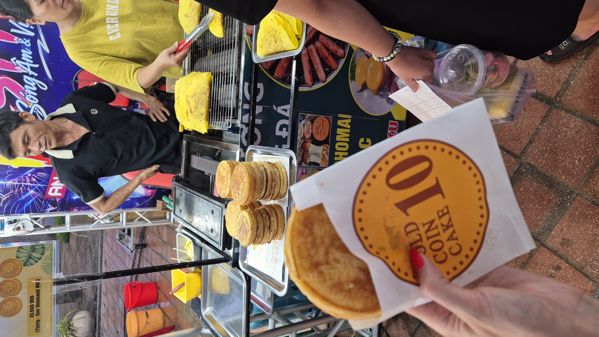 This image captures a vibrant street food scene, likely in Đà Nẵng, Vietnam, where a man is preparing and selling "gold coin cakes." He is wearing a black polo shirt and appears focused on his work. To his right, a woman in a yellow shirt is also involved in the food preparation, holding a pair of tongs.
The stall is set up with various food items and cooking equipment. There are trays of golden-brown, round cakes stacked neatly, and also some triangular yellow crepes being cooked on a grill. Behind the counter, colorful buckets and containers are visible, suggesting a busy operation. A sign displays "35,000 VND (Thirty-five thousand VND)" and the words "NGON TUY" and "HOMAI C," indicating the price and possibly the name of the dish or establishment. Another sign features a graphic of gold coins and the text "GOLD COIN CAKE."
In the foreground, a person's hand, adorned with a bright red nail polish, is holding a piece of paper with the number "10" prominently displayed in a circular emblem, further reinforcing the "gold coin cake" theme. The overall atmosphere seems lively and casual, typical of a bustling outdoor market or street food vendor. The ground is paved with reddish-brown tiles, and the background shows elements of signage and possibly other stalls. The image appears to be taken during the daytime, with natural light illuminating the scene.