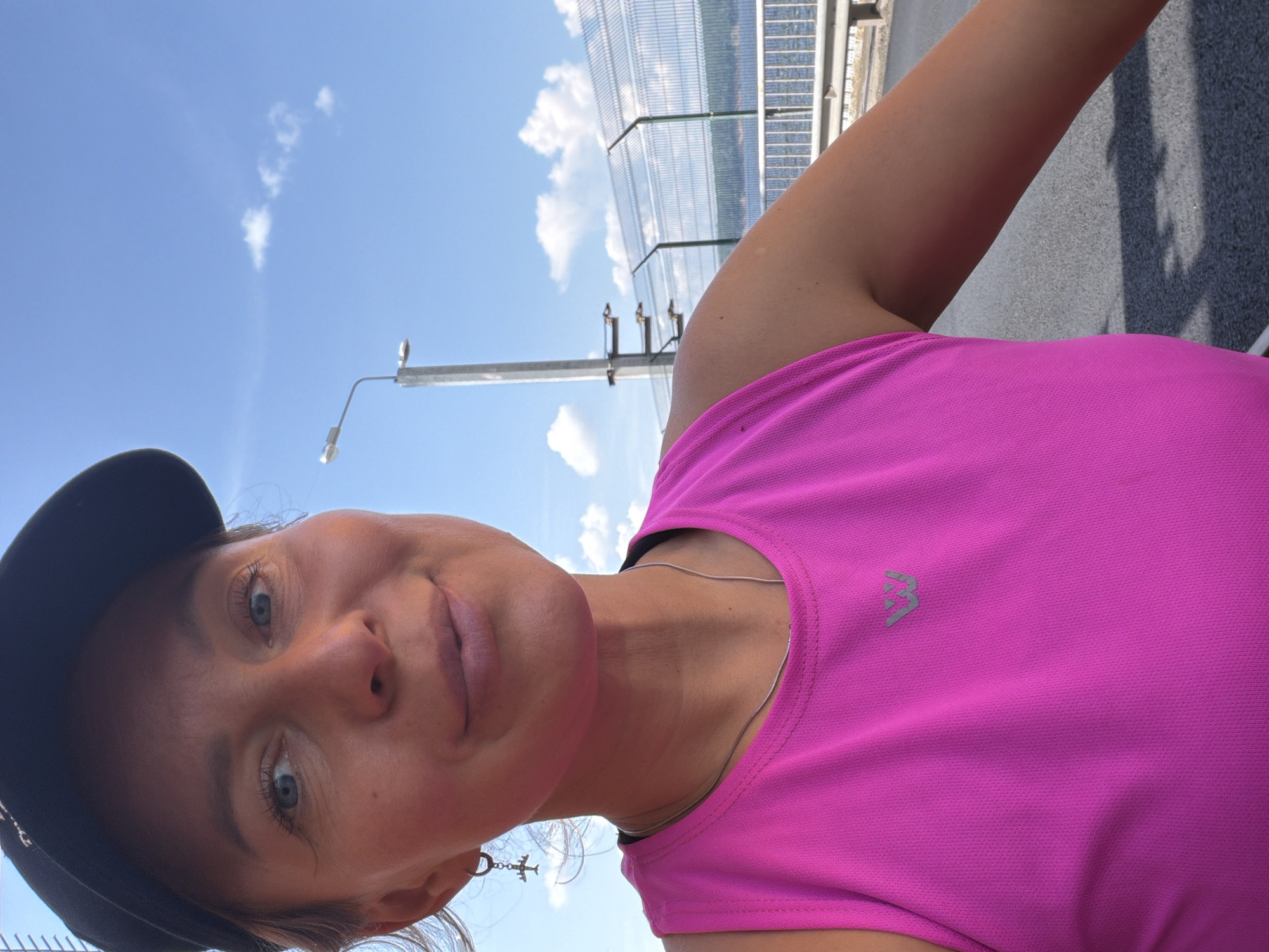 In a close-up selfie taken outdoors, a woman wearing a black baseball cap and a bright pink tank top smiles at the camera. Her light blue eyes are visible, as are her lips, which are slightly parted. She has a thin silver chain around her neck, and a dangling earring in the shape of an airplane on her left ear. The background shows a clear blue sky with a few scattered white clouds, a lamppost, a metal fence, and a body of water, likely the Kama River. The context of Chaikovsky, Russia, suggests this location could be a bridge or embankment along the river in the city. The woman appears to be in good health and spirits, possibly enjoying an outdoor activity on a sunny day.