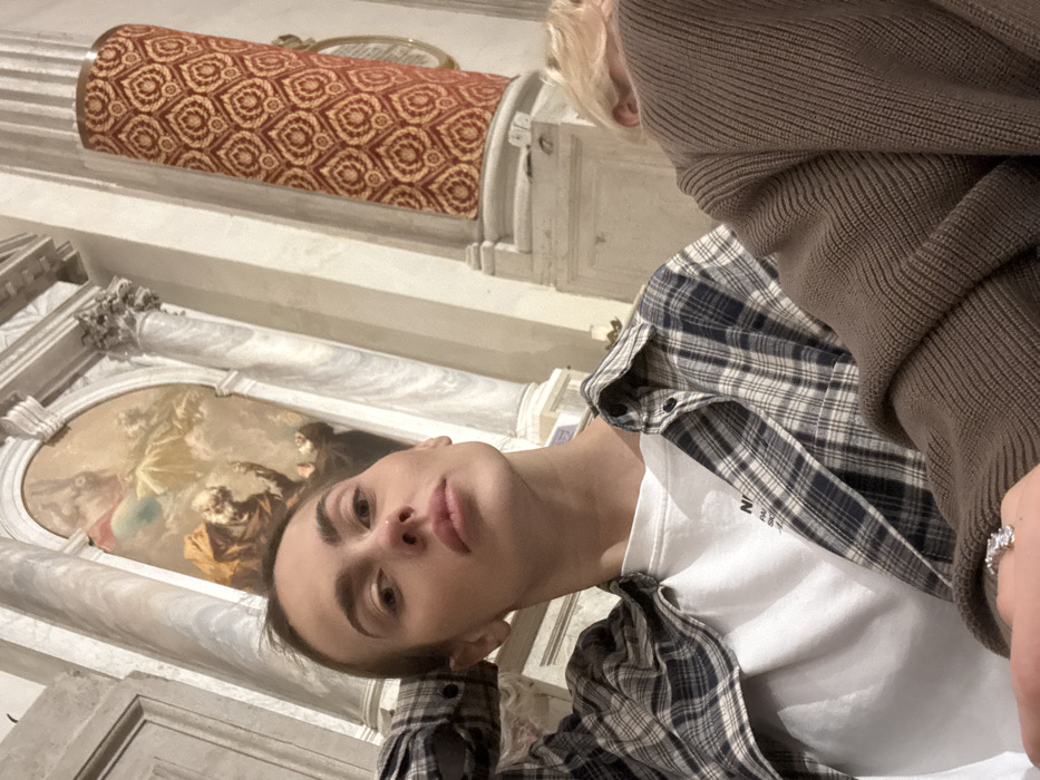 Two individuals are situated inside the Basilica dei Santi Giovanni e Paolo in Venice, Italy, a grand and ornate religious edifice.
In the foreground, a person with light brown hair pulled back, wearing a white t-shirt under an open blue and white plaid shirt, looks directly at the camera with a neutral expression. Their left arm is bent, with the hand resting behind their head/neck. A ring with a clear stone is visible on their left ring finger. To their right, partially obscured, is another person with light-colored hair, wearing a brown ribbed knit sweater. Only the back of their head and shoulder are visible, as they face away from the camera.
The background features classical architectural elements of the basilica. Prominent fluted white marble columns with ornate capitals flank a large, arched altarpiece. This painting, located within the arched frame, depicts a dramatic religious scene, possibly a martyrdom or vision, with figures in the lower portion reaching or looking upwards towards celestial figures in the upper part, characteristic of Italian Baroque artwork. To the right of the painting, another large column is partially visible, covered in a distinctive crimson fabric with an intricate gold and beige patterned motif, likely broccatello. The lighting throughout the interior appears dim, consistent with an historic church setting.
