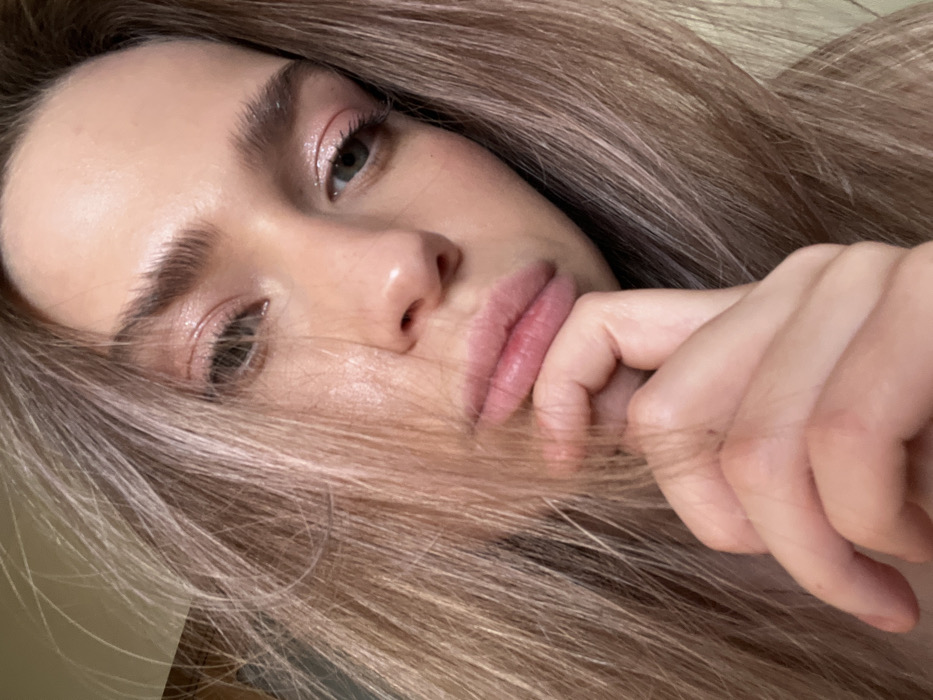 The image displays a close-up, slightly upward-angled portrait of a female subject, estimated to be a young adult. Her light brown hair, appearing somewhat disheveled, frames her face on both sides. Her right hand is partially visible, with the index finger gently pressed against her lower lip and chin area. Her lips are full, natural pink, and slightly parted. Her eyes, appearing light green or hazel, gaze slightly to the viewer's right, exhibiting a glimmer on the corneas. Eyelids are adorned with a shimmering light pink eyeshadow. Her skin has a smooth texture with a slight sheen on the forehead and nose bridge. The background is indistinct, uniformly light-toned, and out of focus, indicating an indoor setting without specific environmental identifiers. The scene is located in Saint Petersburg, Russia.