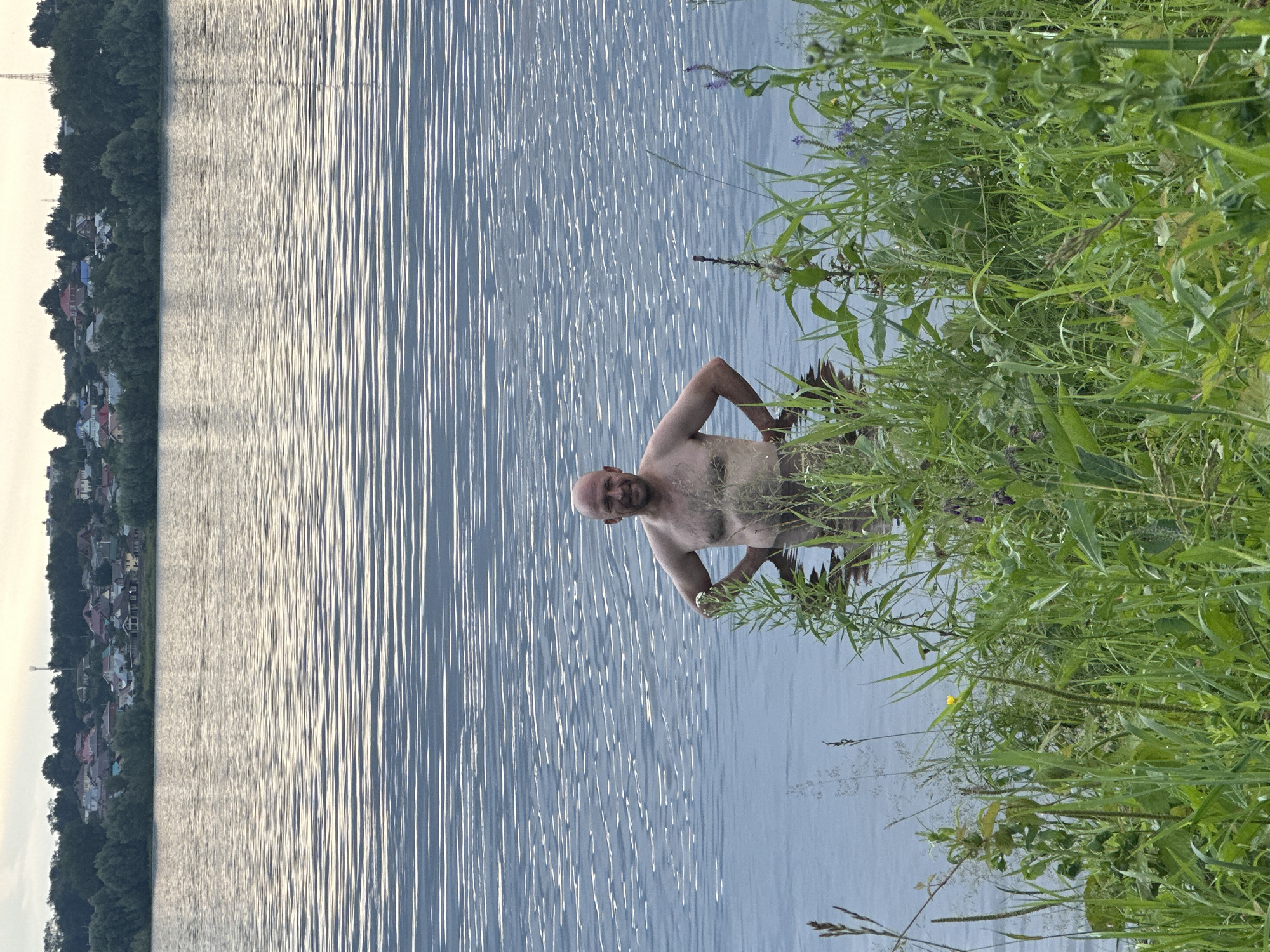 A bald, shirtless man stands in a body of water with his hands on his hips. He is facing the camera and smiling. In the background, there is a shoreline with trees and houses. The water has ripples on its surface, and the sky is overcast. The foreground is filled with tall green grass and some wildflowers. The image appears to have been taken in the city of Noviy, Russia, given the architectural style of the houses and the natural landscape.