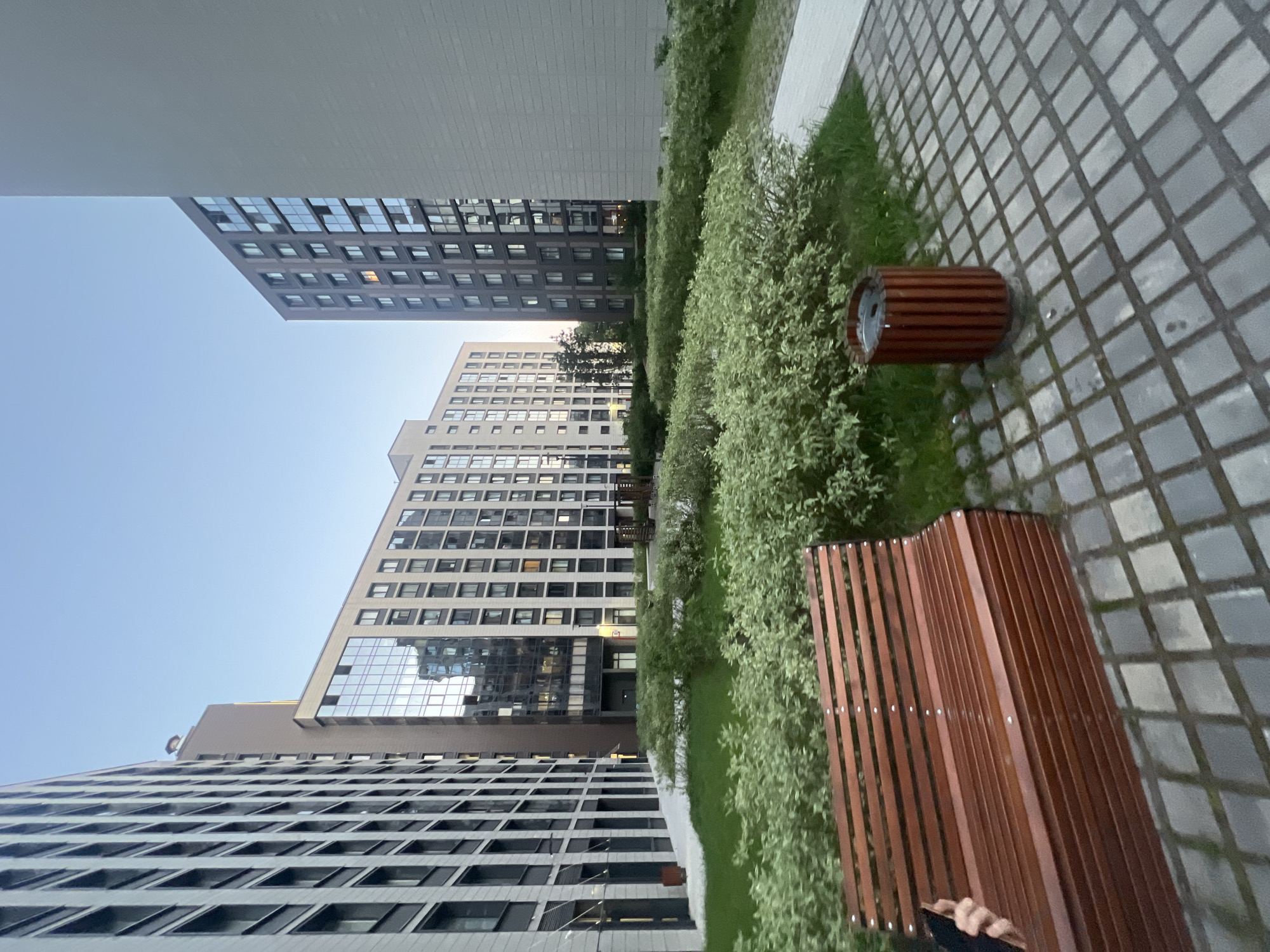 The image depicts an outdoor common area within a modern residential complex in the city of Unknown, Russia, under a clear, pale blue sky. The scene is bordered by several multi-story apartment buildings of varying contemporary designs. To the left, a light-toned building presents a grid facade with dark window frames. In the center, a beige building features a prominent, multi-story reflective glass section, mirroring elements of the surroundings. Further right, a dark grey building with multiple windows, one showing an orange internal light, stands opposite a large, unadorned light grey building wall. The central ground area is a landscaped section with neatly trimmed green grass and clusters of low, variegated green bushes. In the immediate foreground, a dark brown, slatted wooden bench with a curved backrest occupies a paved area consisting of grey rectangular blocks. A human hand, holding a dark smartphone, is visible on the left side of the bench. Adjacent to the bench on the right, a cylindrical wooden trash receptacle, similar in material to the bench, rests on the paving. Paved walkways extend along the base of the left building and into the middle ground on the right.