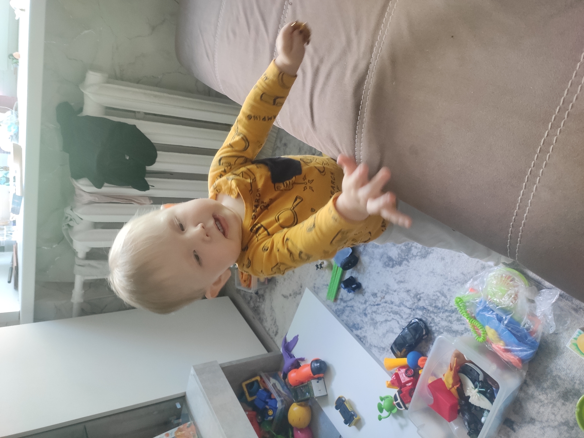 A young child with blonde hair, wearing a yellow long-sleeved shirt with printed graphics, stands near a light-colored sofa and a white storage unit. The child's right arm is raised. Various toys are scattered on a gray, textured floor. The scene includes a white radiator and a black garment hanging on it. The location is possibly in Sterlitamak, Russia.