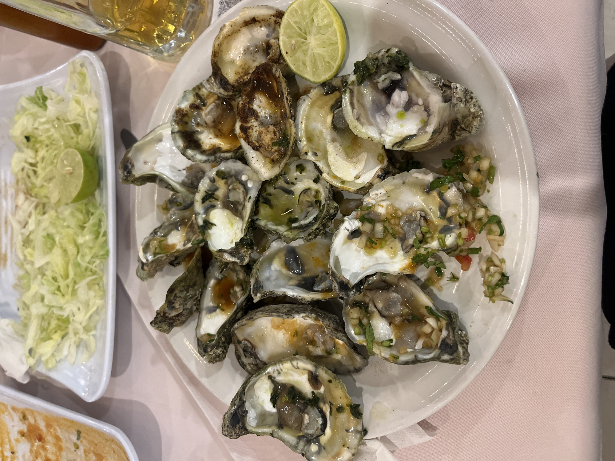 The image shows a plate of oysters on a light pink surface, likely a tablecloth. The oysters are arranged in a heap and appear to be prepared with different toppings or sauces. A lime wedge sits atop the oysters. To the left of the oyster plate, there's a container of shredded lettuce with another lime wedge. A glass bottle, possibly containing a beverage, is visible in the upper left corner. Another container with food that appears to be a baked dish is partially visible in the lower left corner. The photo was taken in the city of San Bernardino Tlaxcalancingo, Mexico.