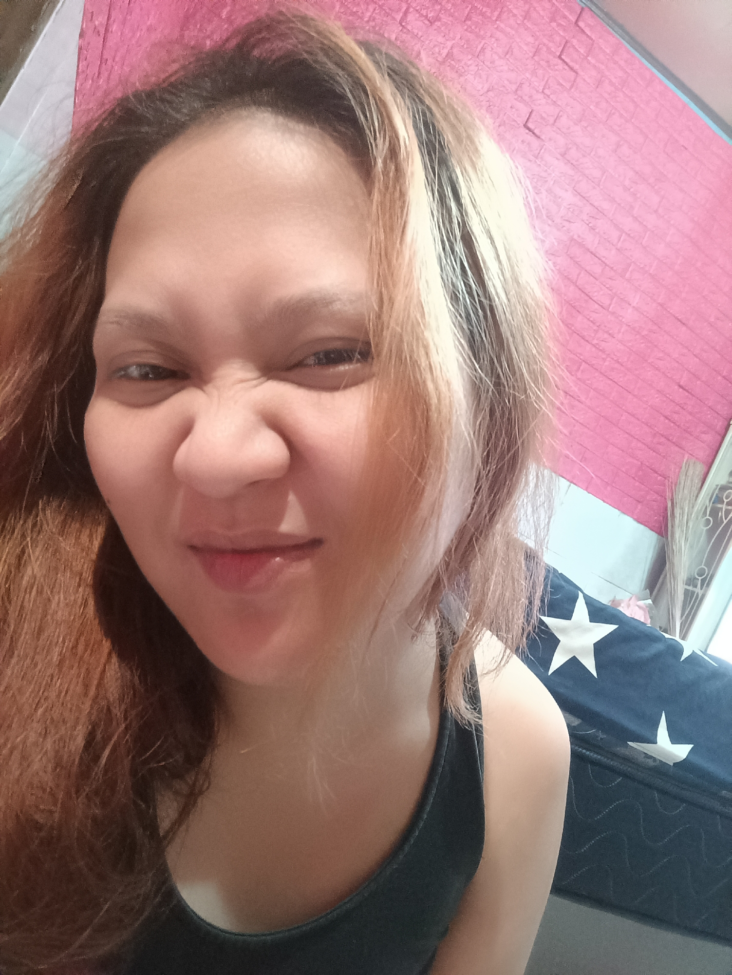 A medium-shot selfie shows a person making a grimacing face. The person has reddish-brown hair that is lightened in some areas. The person is wearing a dark tank top. In the background, there is a pink textured surface, a blue mattress with white stars, and a window with blinds. The overall scene appears to be an indoor setting, likely a bedroom.