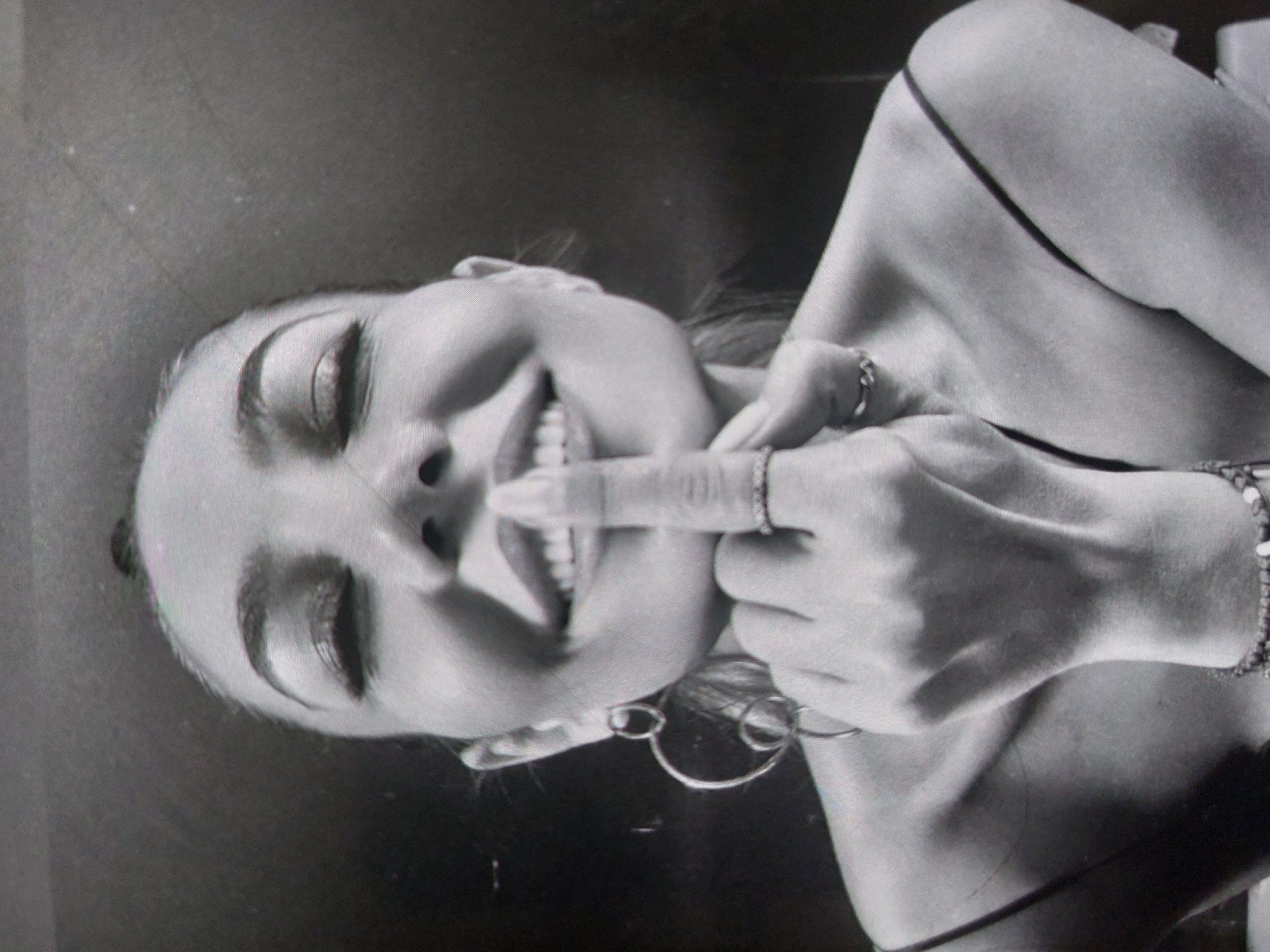 The image is a black and white close-up of a woman smiling and holding her finger to her lips. She is wearing hoop earrings, a ring on her finger, and a bracelet. The background is dark and out of focus. There is no visible information about the location in the city of Tsarevo, Bulgaria.