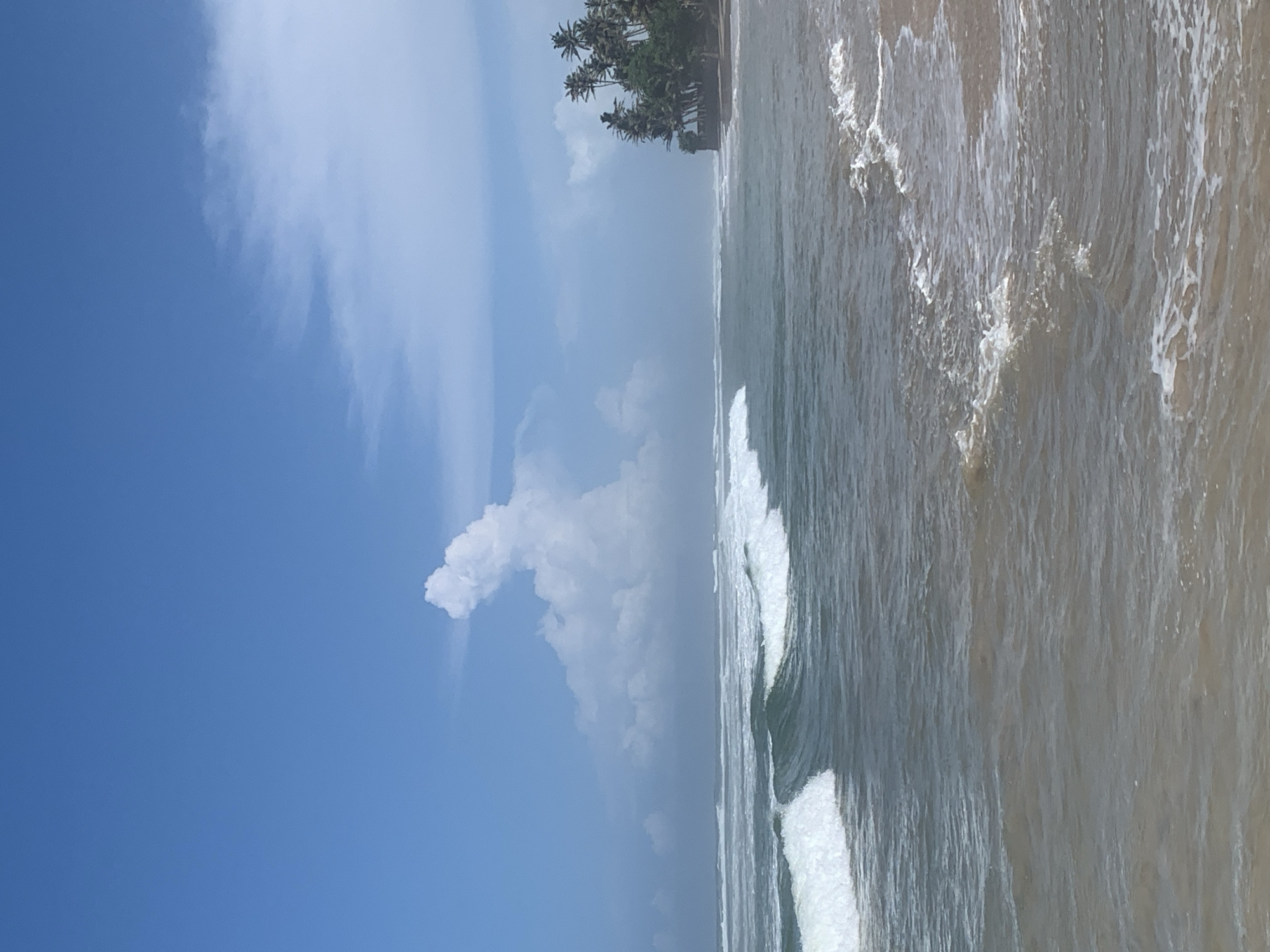 This image captures a coastal scene in Kathaluwa, Sri Lanka. The sky is predominantly blue with wispy clouds. The ocean waves, white with foam, crash onto a sandy beach. Palm trees line the shoreline in the distance.