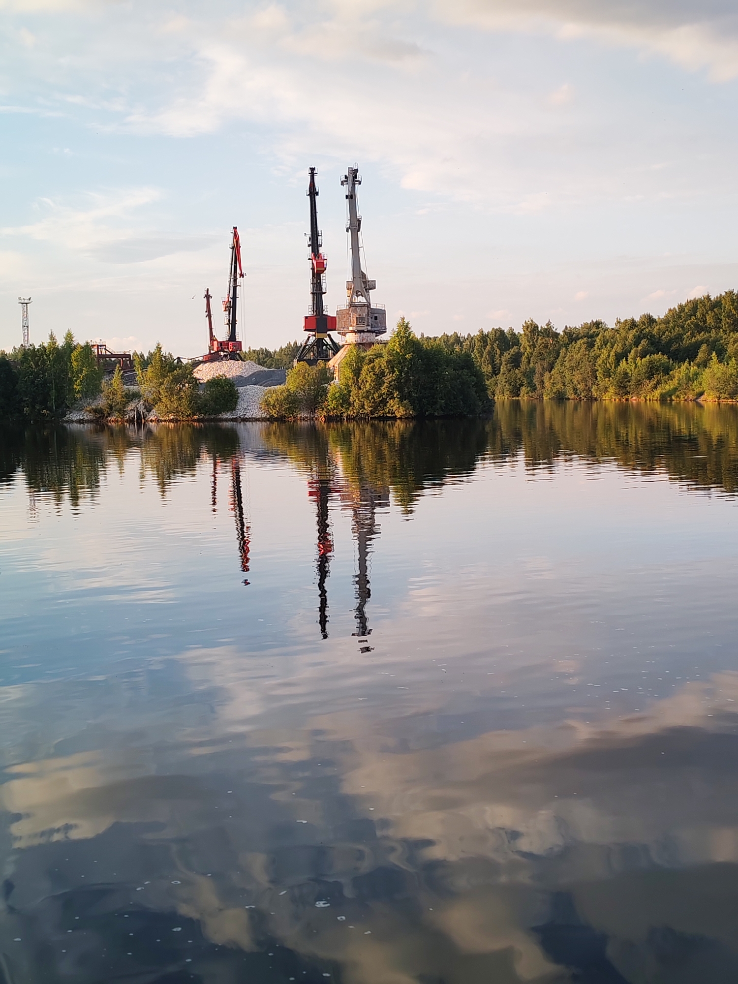 The image captures a serene water scene with industrial elements on the far bank. Three large cranes, two red and white and one grey and white, stand prominently against a light blue sky with scattered clouds. They are situated behind a mound of gravel, surrounded by lush green trees. The water in the foreground reflects the sky and the cranes, creating a symmetrical and tranquil effect. The scene appears to be a quarry or a port area on the outskirts of Aleksandrovskoe, Russia, given the industrial machinery and the natural landscape. There are no people visible in the image, and the overall atmosphere is one of quiet industrial activity and natural beauty.