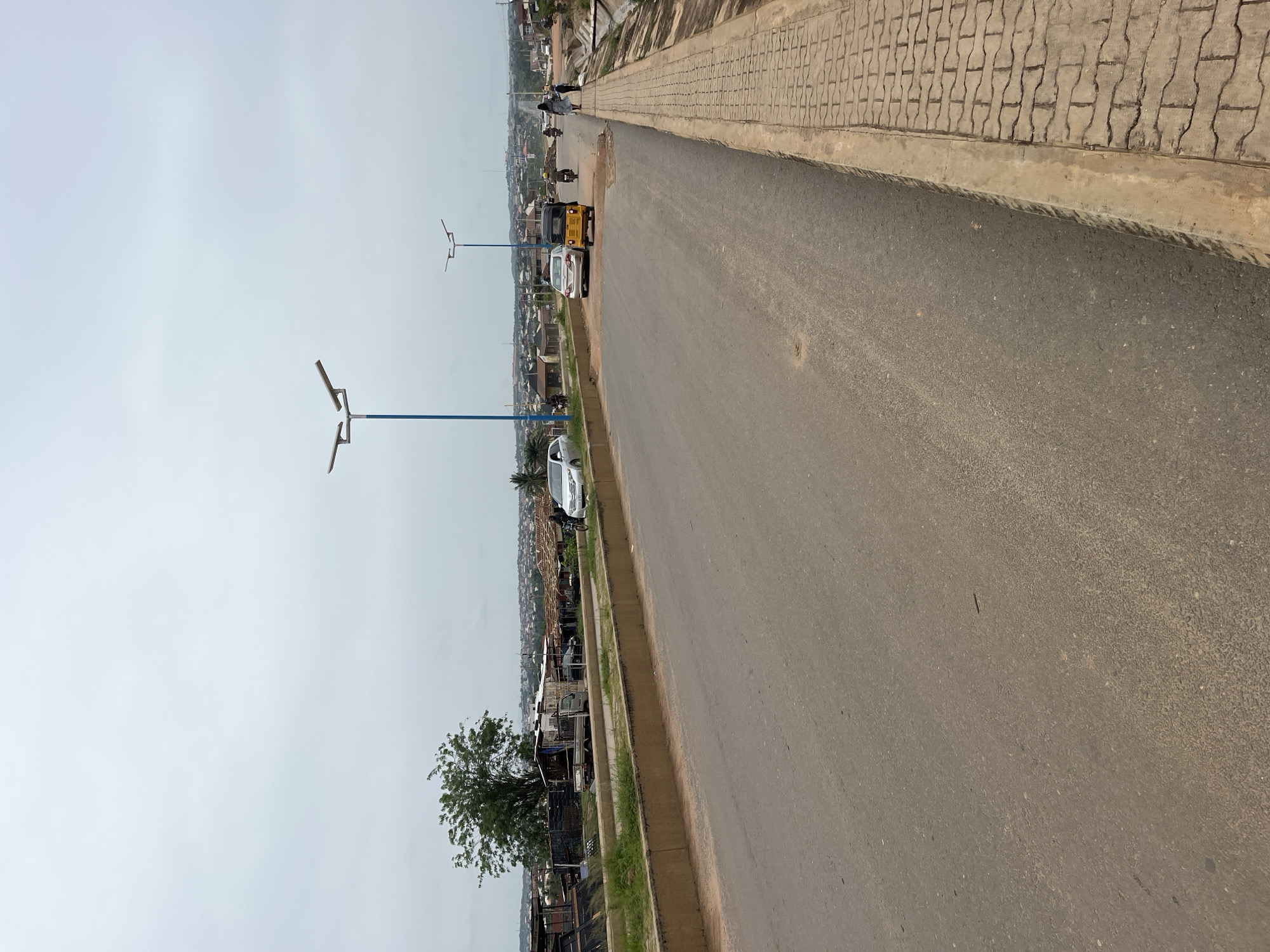 The image depicts a view of a street in Osogbo, Nigeria. The street is asphalt and has a light-colored sidewalk made of interlocking paving stones. A blue light pole stands on the left side of the road. The street has vehicles parked on the left-hand shoulder. A tree and several stalls or shops can be seen on the left, just off the road. In the distance, the road extends into the cityscape with houses and buildings visible. Some people are walking on the sidewalk in the distance. The sky is overcast.