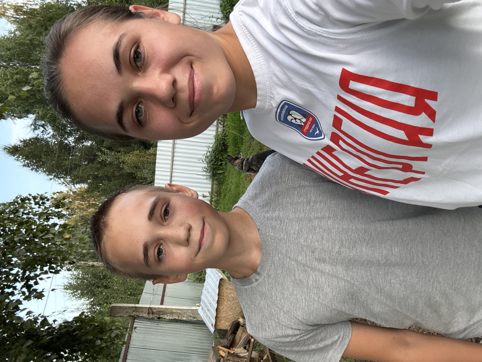 Two people, a boy and a girl, stand in front of a fence in a rural setting, possibly the city of Elovo, Russia, given the presence of birch trees and a modest structure. The boy on the left, wearing a grey t-shirt, has short brown hair and a light complexion with freckles. He is looking towards the camera with a slight smile. The girl on the right, wearing a white t-shirt with red lettering and a circular emblem, has brown hair pulled back and a warm smile. Her t-shirt bears the inscription "СТУДЕНЧЕСКАЯ ЛИГА" (Student League) and what appears to be a sports logo. Behind them, the environment includes a metal fence, stacks of firewood, a small wooden structure with a corrugated metal roof, lush green grass, and dense trees in the background. The lighting suggests it is daytime, with sunlight casting shadows. A dog can be seen in the middle ground, between the two individuals and the fence.