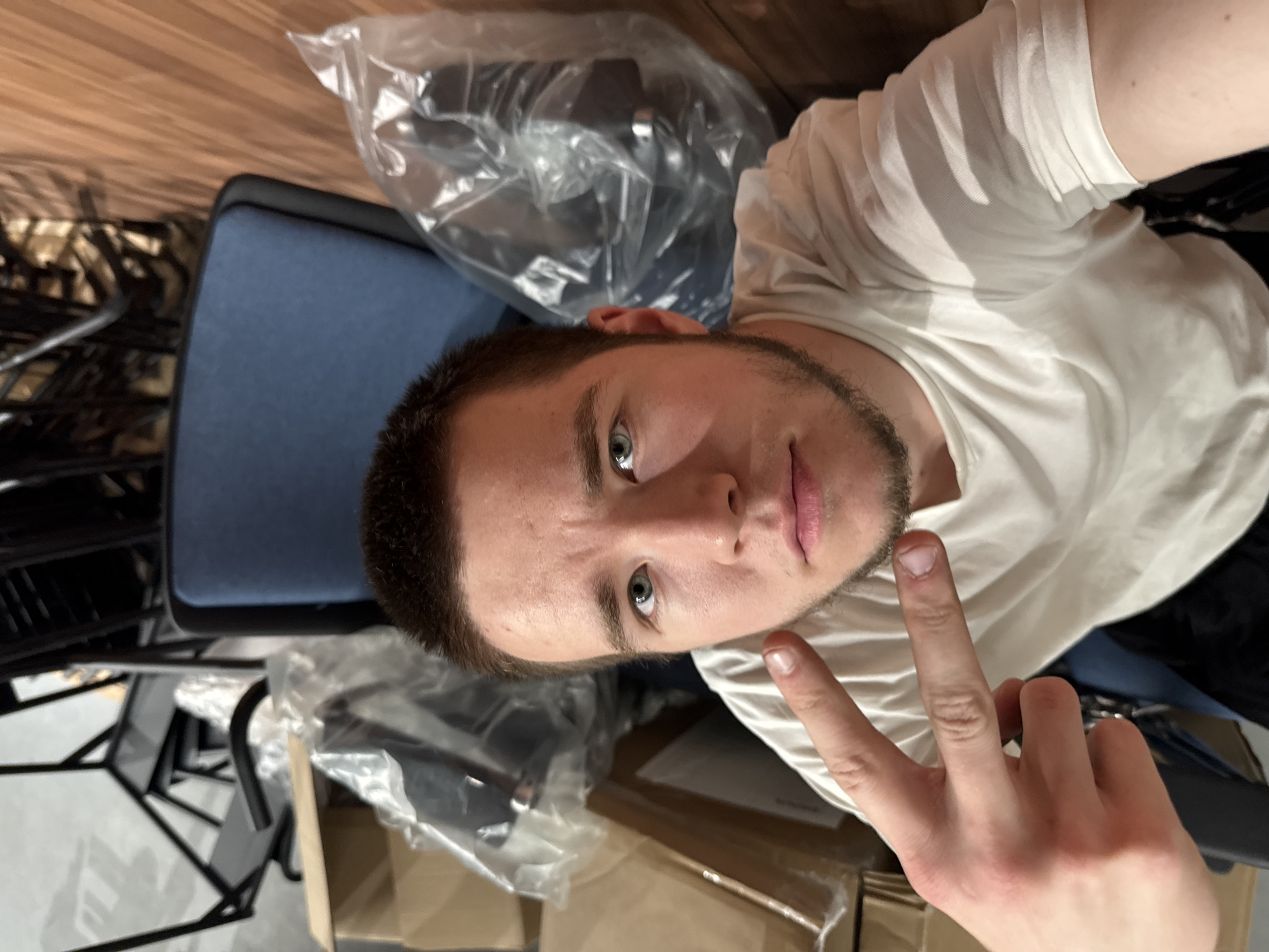 A young man is taking a selfie. He has short brown hair, light eyes, and a trimmed beard. He is wearing a white t-shirt and is making a peace sign with his right hand. The background includes a partially assembled chair with a blue seat, packing materials, and wood flooring. The image provides no verifiable information to indicate the location is in Kazan, Russia.