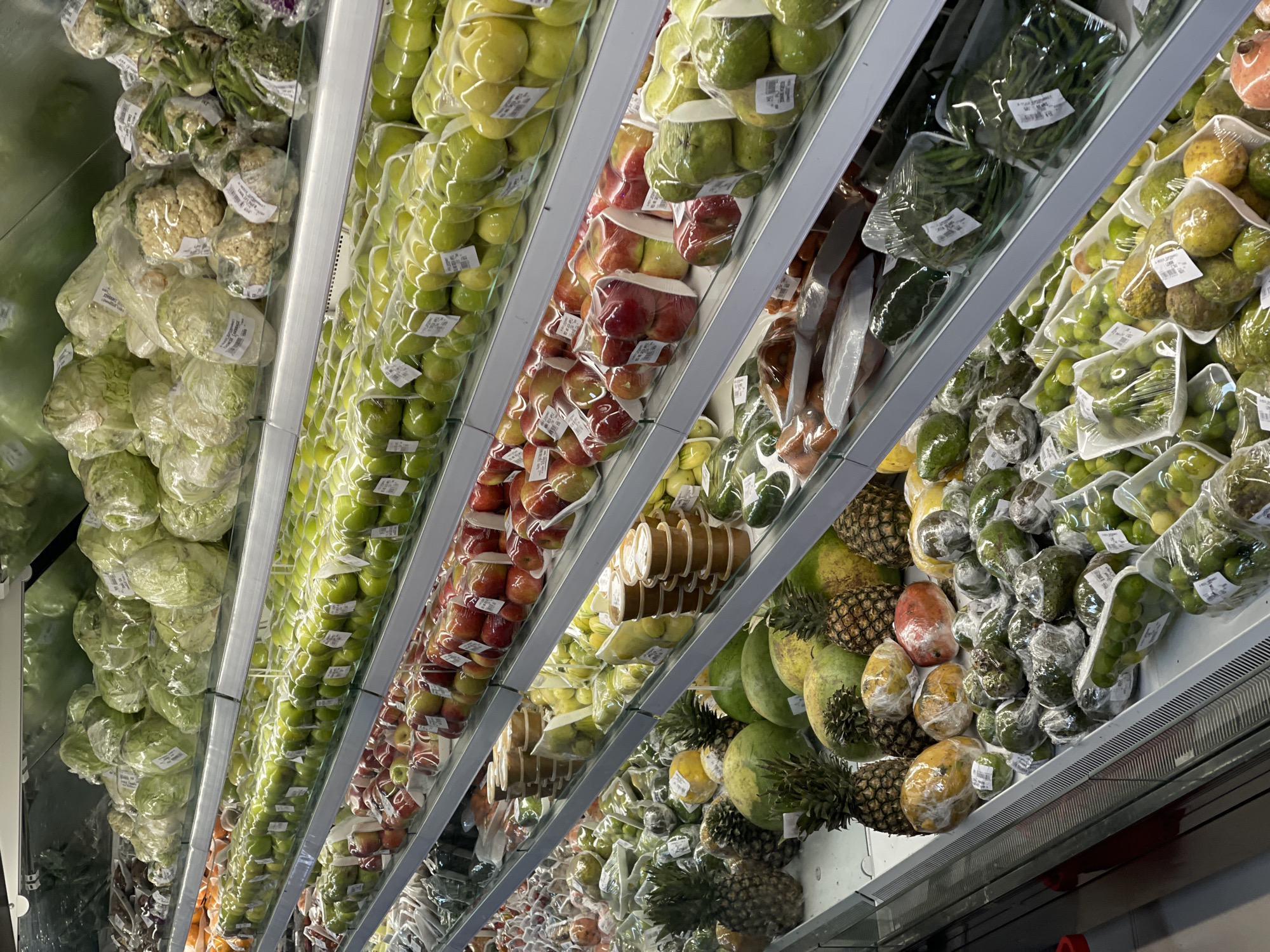 A wide-angle, eye-level shot displays rows of fresh produce arranged on refrigerated shelves in a supermarket, likely located in Guzape, Nigeria. The foreground is dominated by an assortment of fruits including pineapples, papayas, guavas, and limes, many of which are individually wrapped in plastic. Further back, shelves are stocked with green apples, red apples, cauliflowers, cabbages, and what appear to be bunches of green beans. The lighting is bright and even, typical of a retail environment, illuminating the vibrant colors of the fruits and vegetables. The shelves are made of metal and glass, with refrigeration units visible along the bottom of the display. There are no people visible in the image, and the focus is entirely on the produce. The overall impression is one of abundance and organization within a commercial setting.