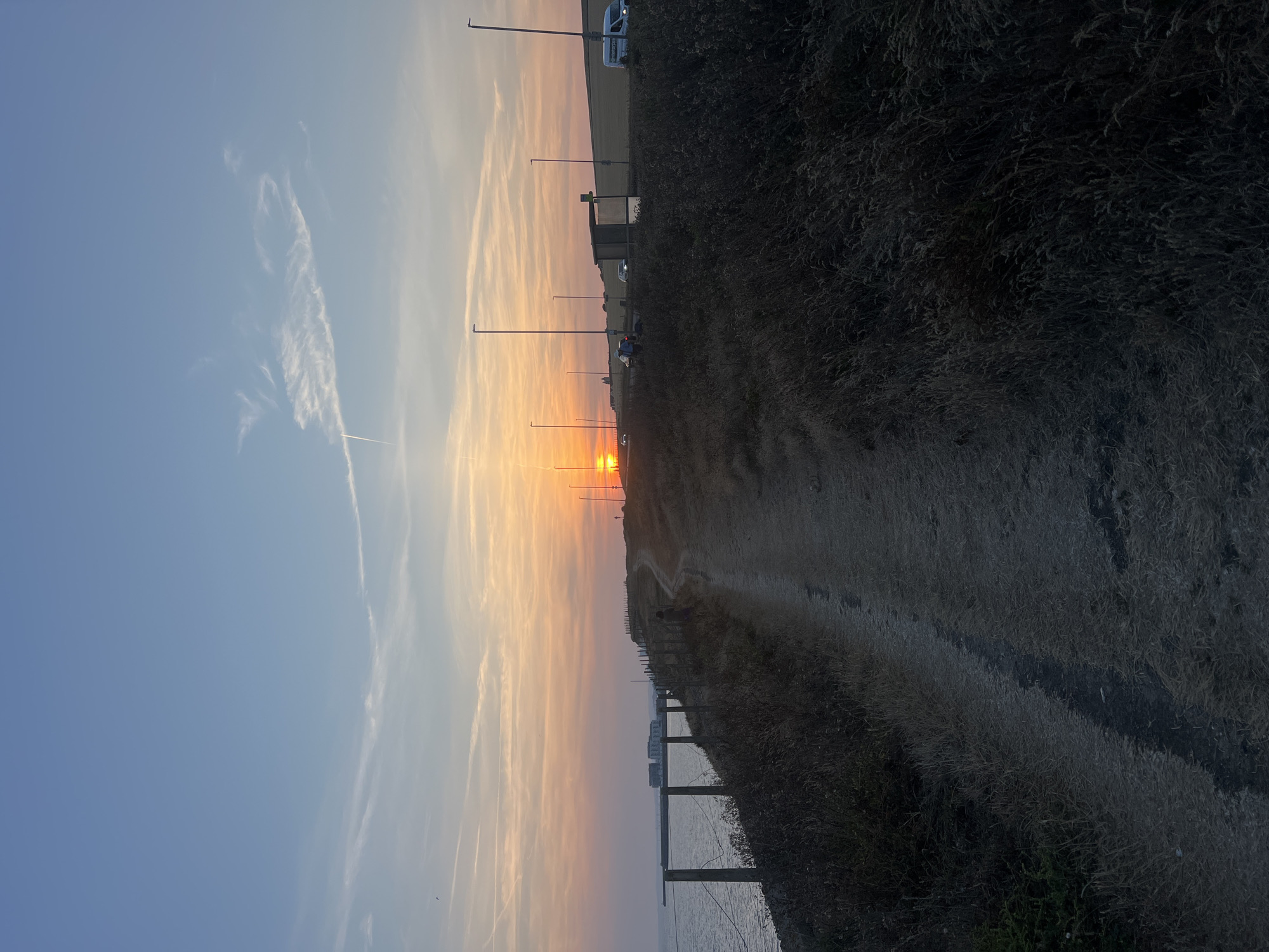 The image depicts a coastal path at sunset in Ovingdean, United Kingdom. The sun is low on the horizon, casting a warm glow across the sky and illuminating thin, wispy clouds. The sea is visible to the left, with a fence line running along the edge of the path. The path itself is dirt and winds uphill. On the right side of the path, there is dense, dry grass and a road with streetlights and a few vehicles, including a white van and a bus stop shelter. In the distance, there are some buildings, possibly residential, and a field behind the road. A single person is visible walking on the path. The overall scene is tranquil and evokes a sense of end of day.