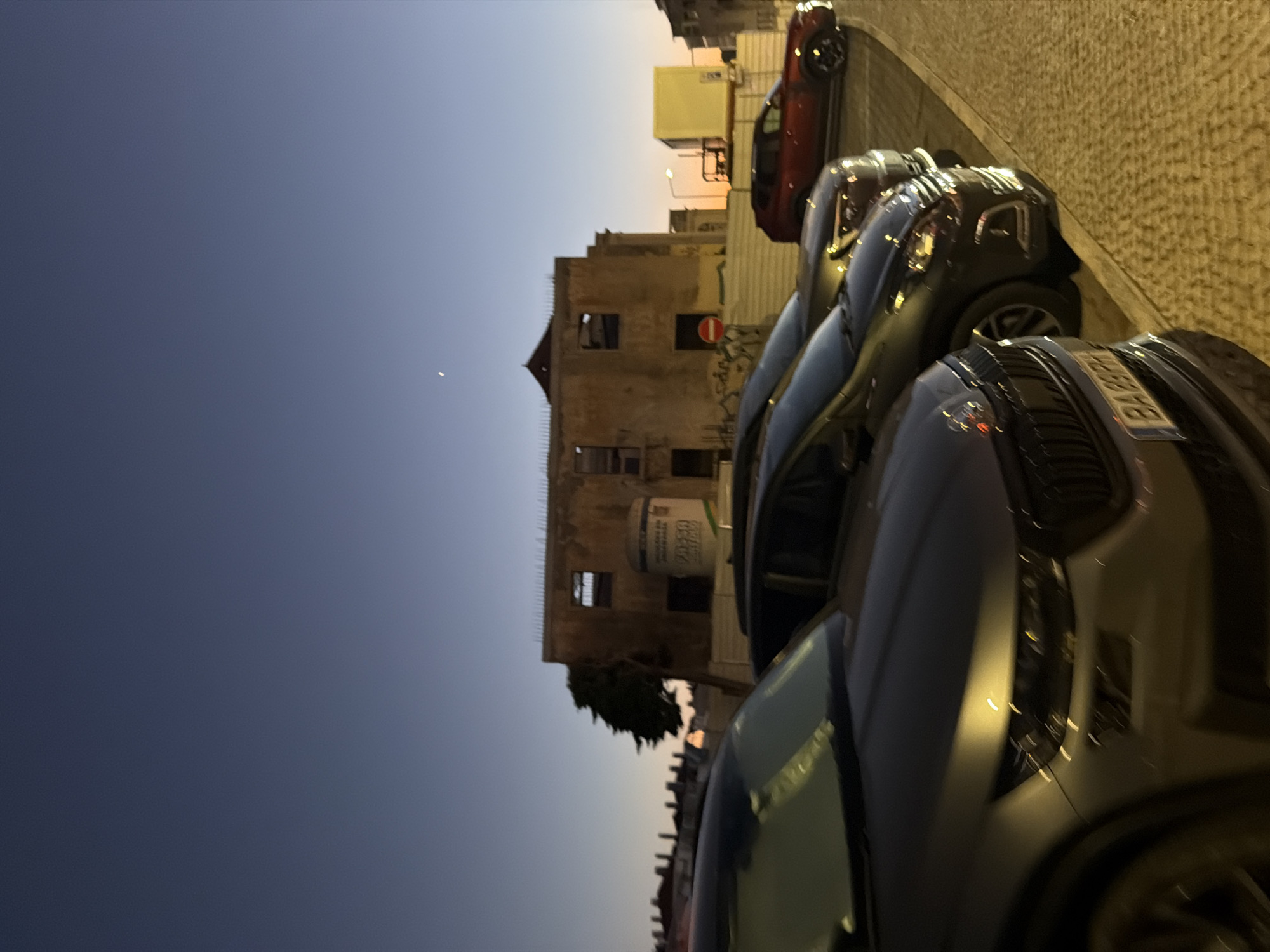 The media file depicts a street scene in Cascais, Portugal, likely during dusk. The primary subjects are several cars parked along a cobblestone street. The cars are of various colors, including dark grey, black, and a red one further in the background. The setting appears to be urban, with buildings visible on the right side of the frame. One building is an older, possibly abandoned structure with visible damage and dark window openings. Another building features what looks like a large white cylindrical container attached to its exterior. The sky transitions from a deep blue to a lighter gradient near the horizon, suggesting either sunrise or sunset. The lighting on the cobblestones indicates artificial streetlights are on. There is a round red sign with a white line on it attached to the older building. The overall mood is quiet and still, characteristic of a transitional time of day. The license plate on the closest car is partially visible and reads "BA".