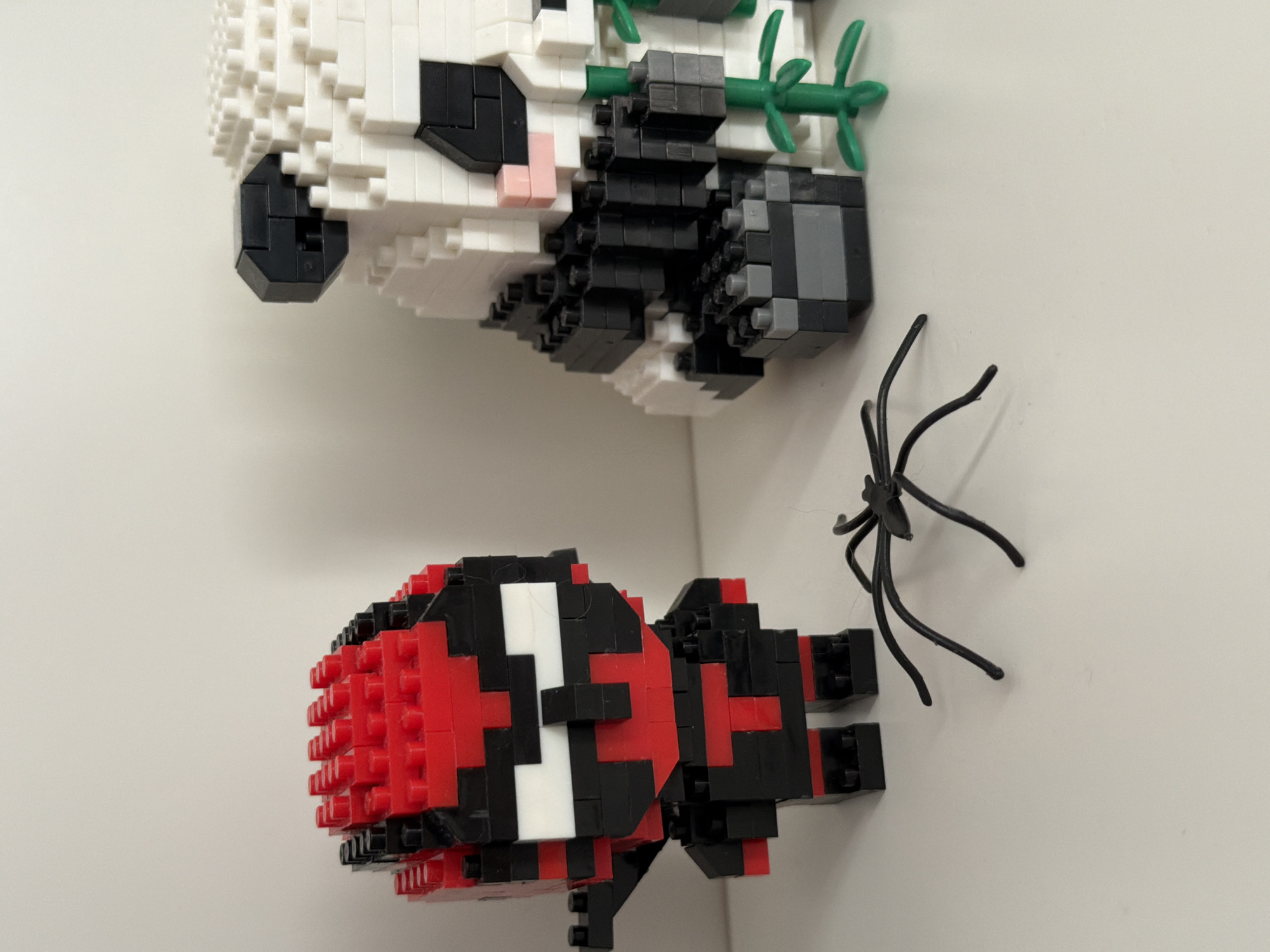 The image shows two brick-built figures, a red and black character resembling a ninja or superhero, and a white and black panda, standing on a white surface. A small black plastic spider is positioned on the surface in front of the figures. The background is a plain, light-colored wall. There is no indication of the location being in Kyiv, Ukraine, or any specific city. The scene appears to be a still life arrangement of toys.