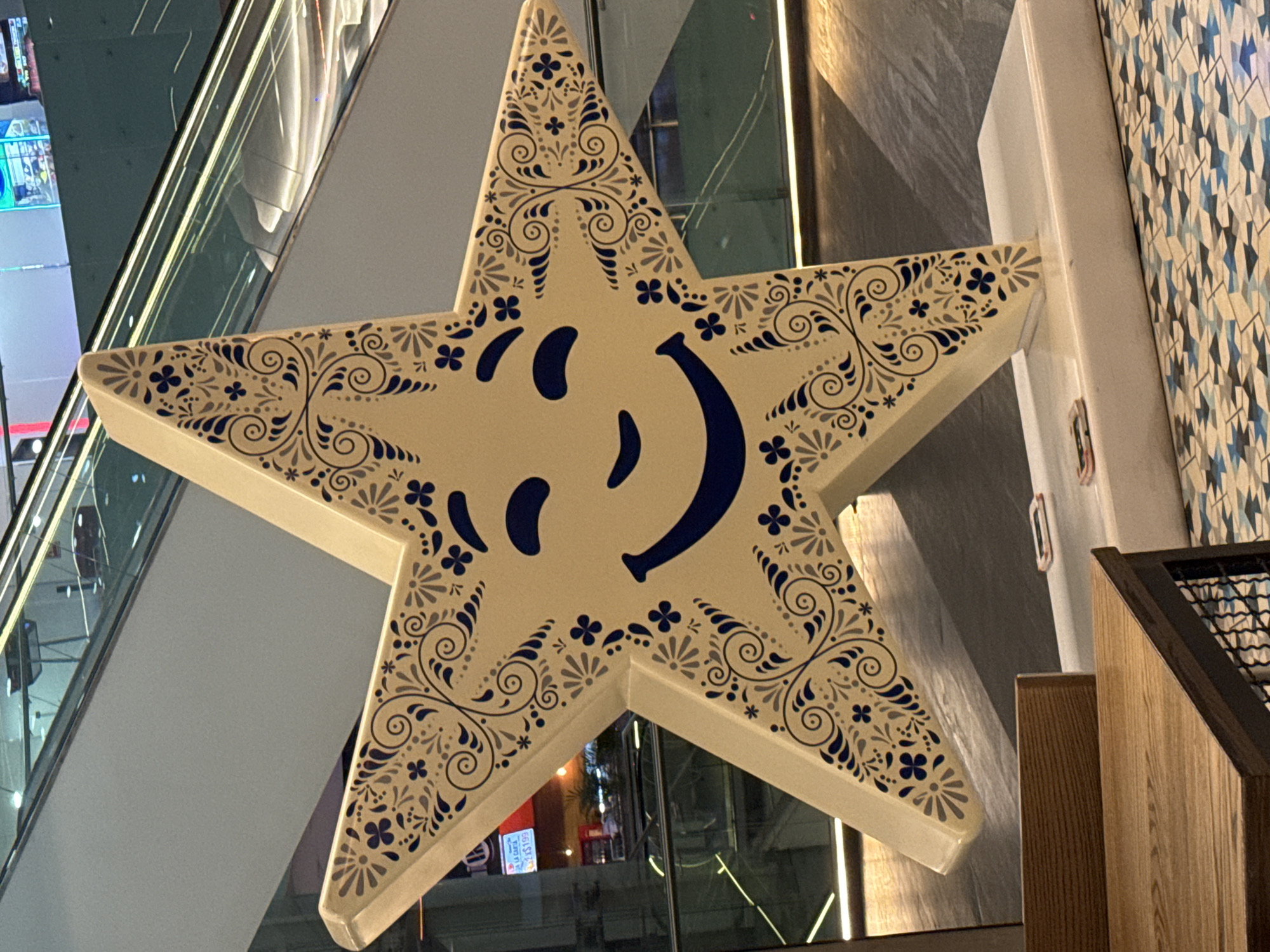 The image shows a large, five-pointed star-shaped sign with a white background and blue decorative patterns. The sign features a stylized smiley face. The star is mounted on a wall with a patterned blue and white design. In the background, there are visible elements of a modern building with an escalator, glass windows, and interior lighting. The scene suggests an indoor location, possibly a shopping mall or commercial space. While the location is stated to be in Puebla, Mexico, there are no visual cues in the image to explicitly confirm this.