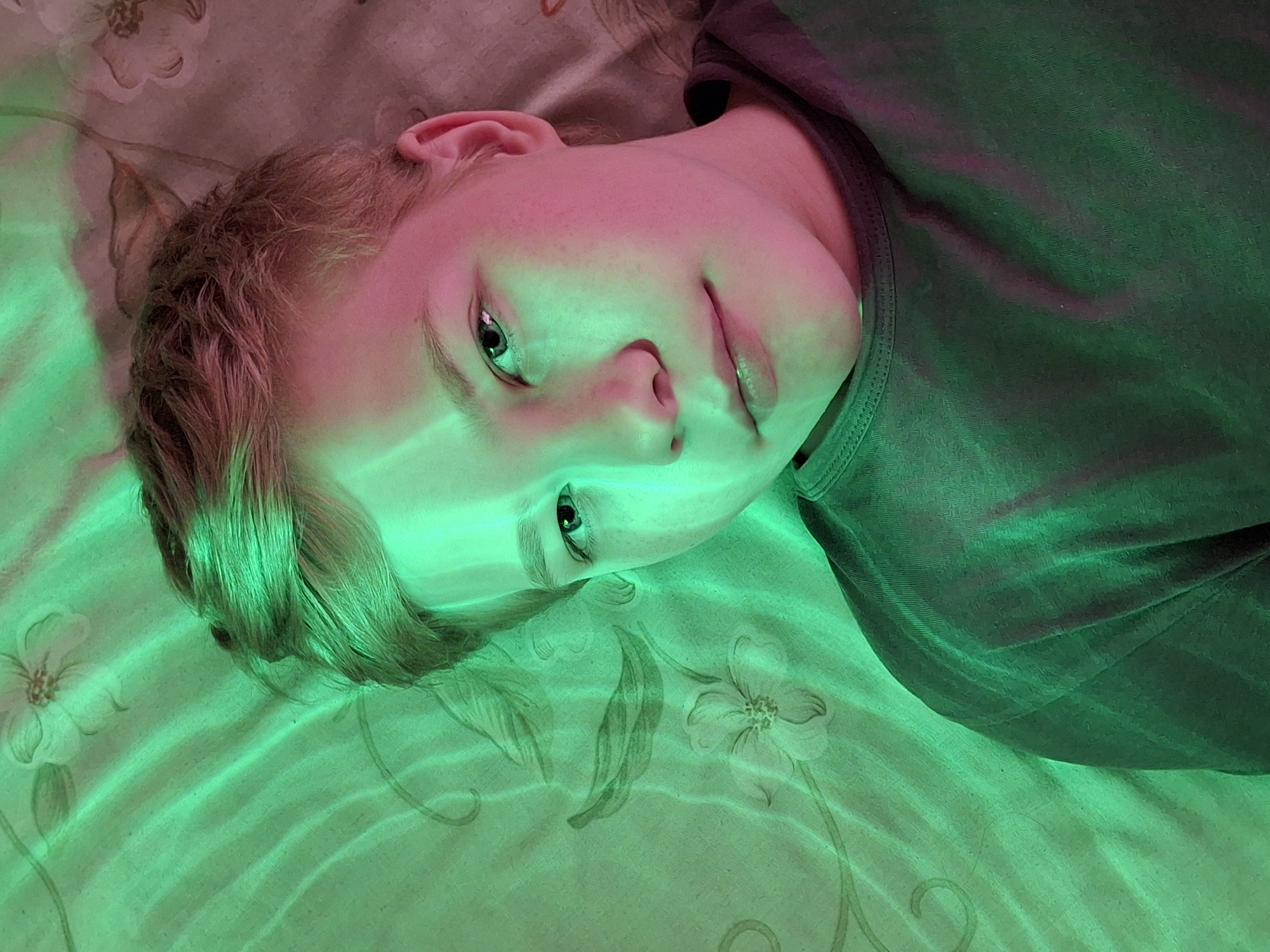 The image shows a close-up, overhead shot of a young person lying down, illuminated by green and pink light. The person has light brown hair and is looking towards the camera with a neutral expression. They are wearing a dark t-shirt. The background appears to be a patterned fabric, possibly a bedsheet or curtain, with faint floral designs. The lighting creates strong shadows and highlights across the person's face and hair. There is no discernible city or location information in the image.