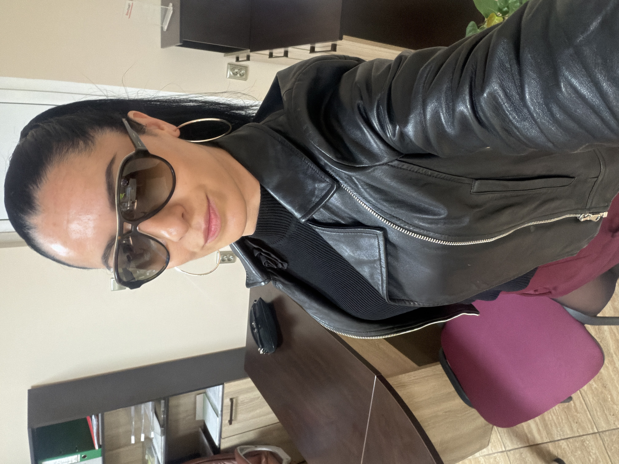 A woman with dark hair pulled back, sunglasses, and gold hoop earrings poses indoors, wearing a black leather jacket over a black top. She is seated in an office or workspace, with a desk visible on the left containing office supplies. A burgundy office chair is to her right. The location is in Pleven, Bulgaria.