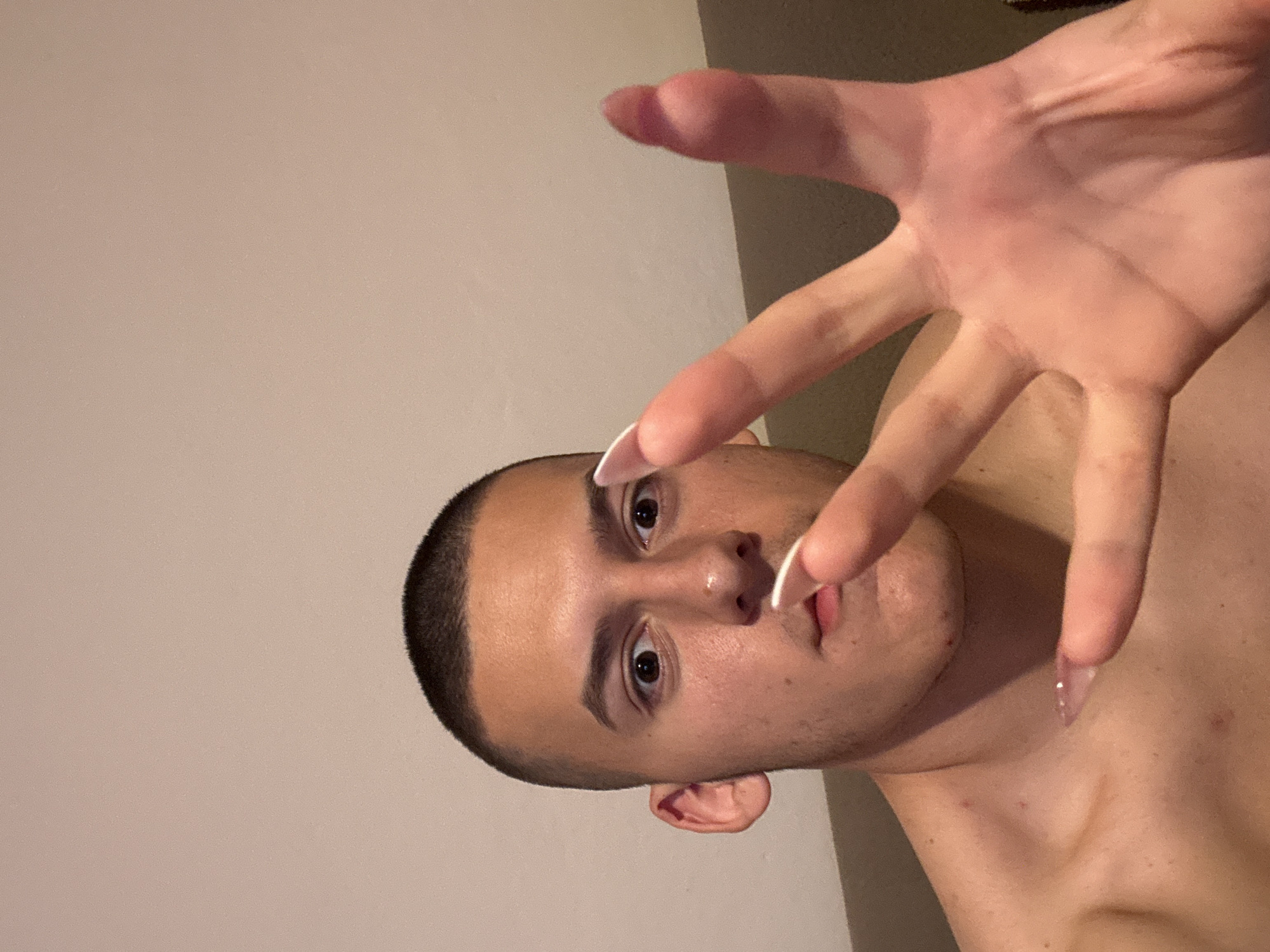 A person with short, dark hair and light skin is posing for a photo. They are holding up their right hand towards the camera, displaying long, artificial nails with a white tip design. The person is shirtless, and the background consists of a beige wall. The location is not discernible from the image.