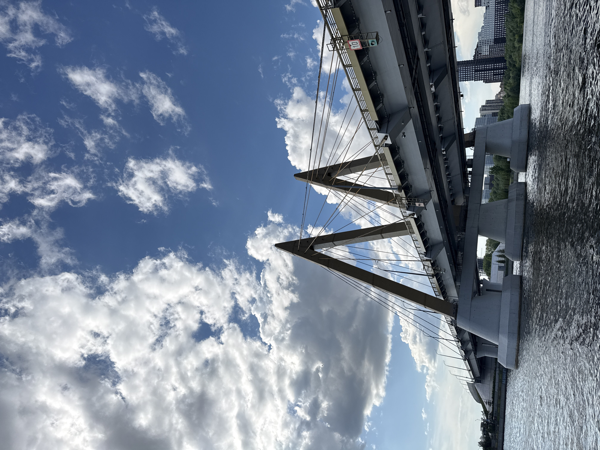 The image depicts the Millennium Bridge in Kazan, Russia, viewed from a low angle. The bridge's structural elements, including its cable-stayed design and support pylons, are prominently featured against a backdrop of a partly cloudy sky. In the distance, buildings are visible. The bridge spans a body of water, with ripples on the surface.