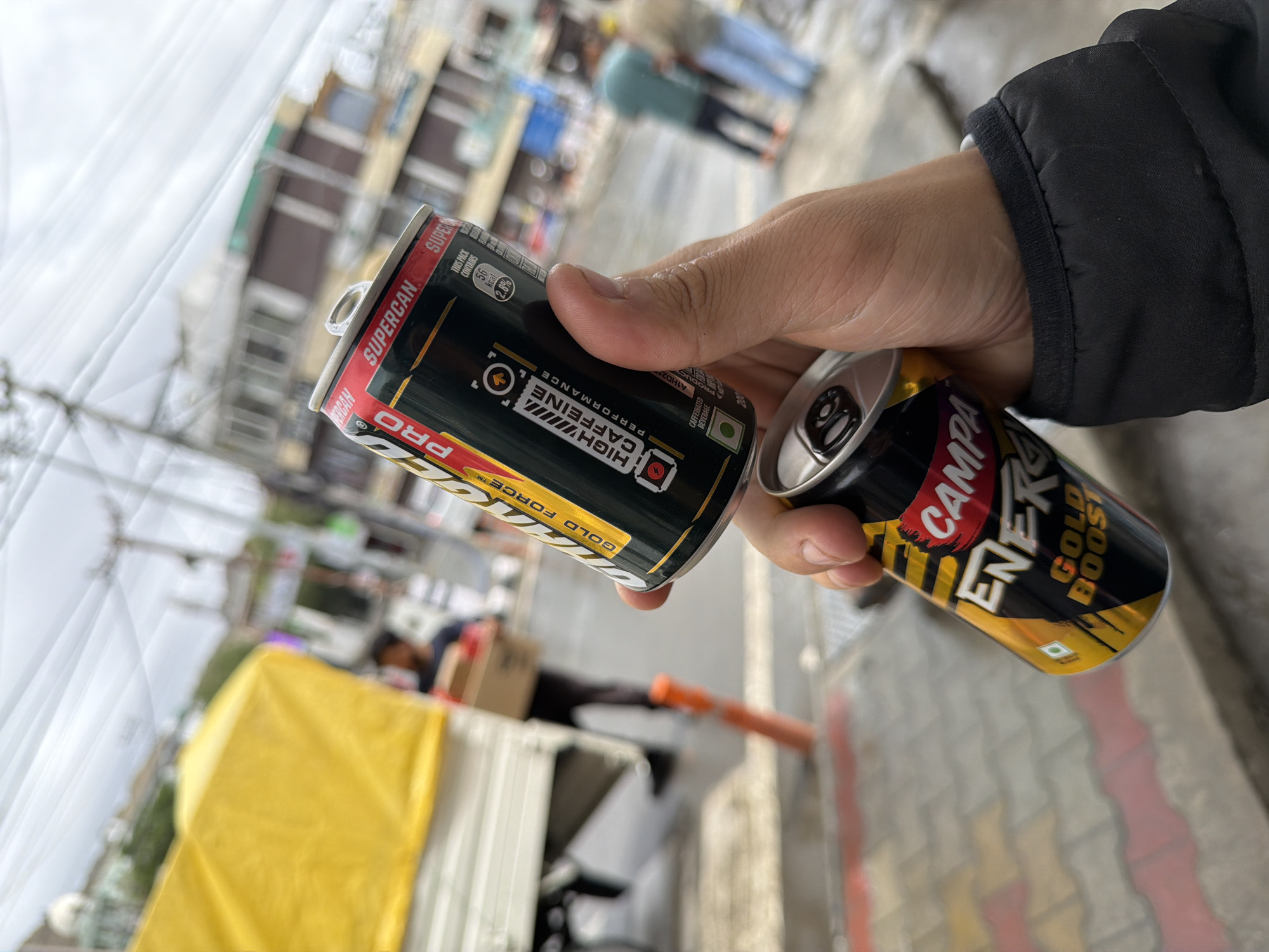 The image shows a person holding two energy drink cans in their hand, in the city of Leh, India. The first can, partially obscured by the second, is a red, black, and gold can labeled "PRO" and "SUPERCAN" with text indicating "HIGH CAFFEINE PERFORMANCE". The second can, closer to the camera and more visible, is a gold and black can labeled "ENERGY" and "GOLD BOOST". The background features a street scene with a yellow-covered truck on the left, some buildings, and a few people walking in the distance. The ground appears wet, suggesting it might have rained. The sky is overcast. The overall impression is a casual moment captured outdoors in an urban environment in Leh.