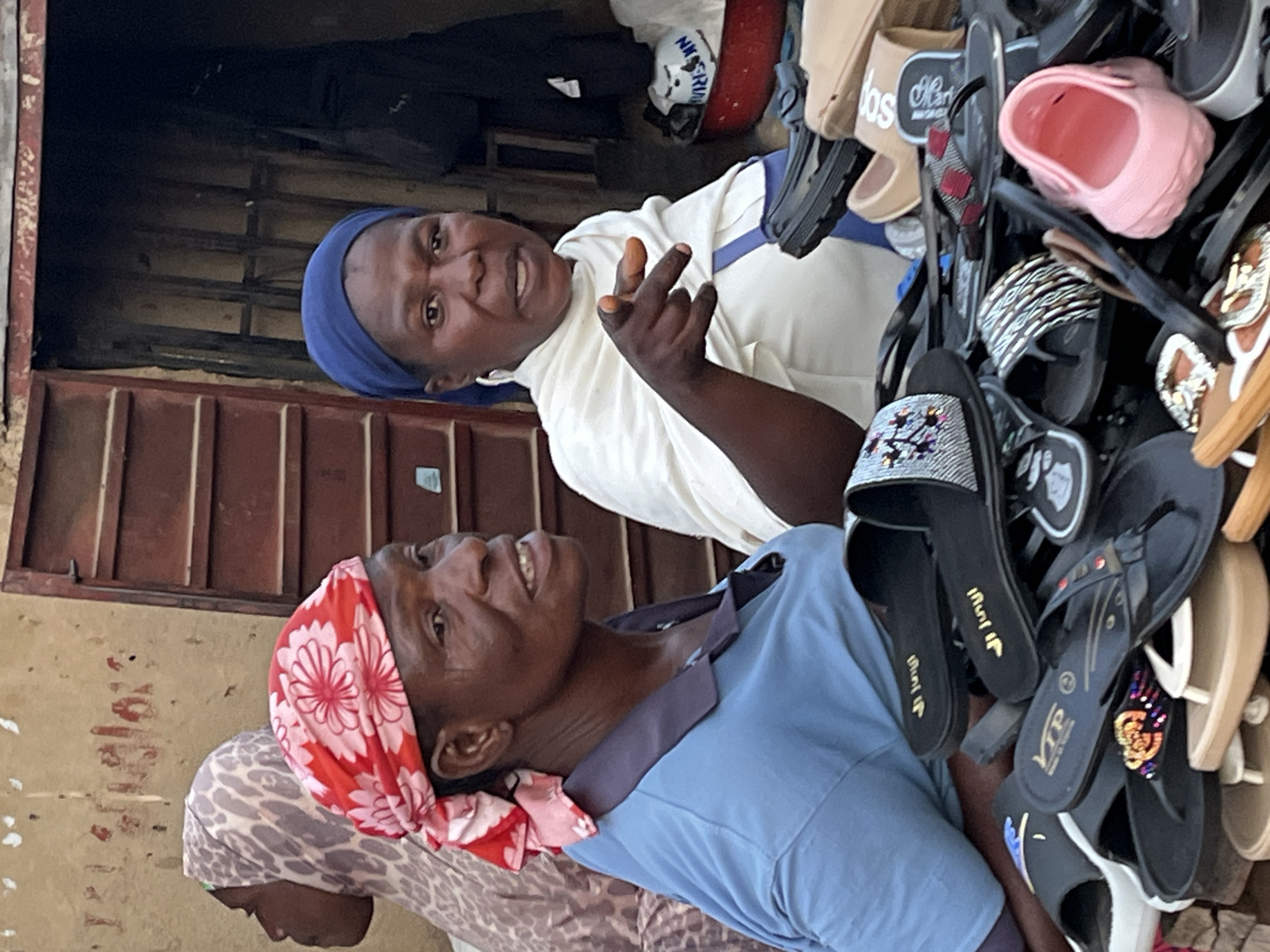 In Jos, Nigeria, a street vendor is seen with a display of sandals for sale. She is wearing a blue polo shirt and a red and pink floral headscarf. Another woman to her right wears a blue headscarf and a white garment, gesturing with her right index finger. Behind them, a third person in a patterned headscarf is partially visible. The background features a rustic stall with a brown metal door. The ground is covered with a variety of sandals, including flip-flops and slides, in assorted colors and designs.