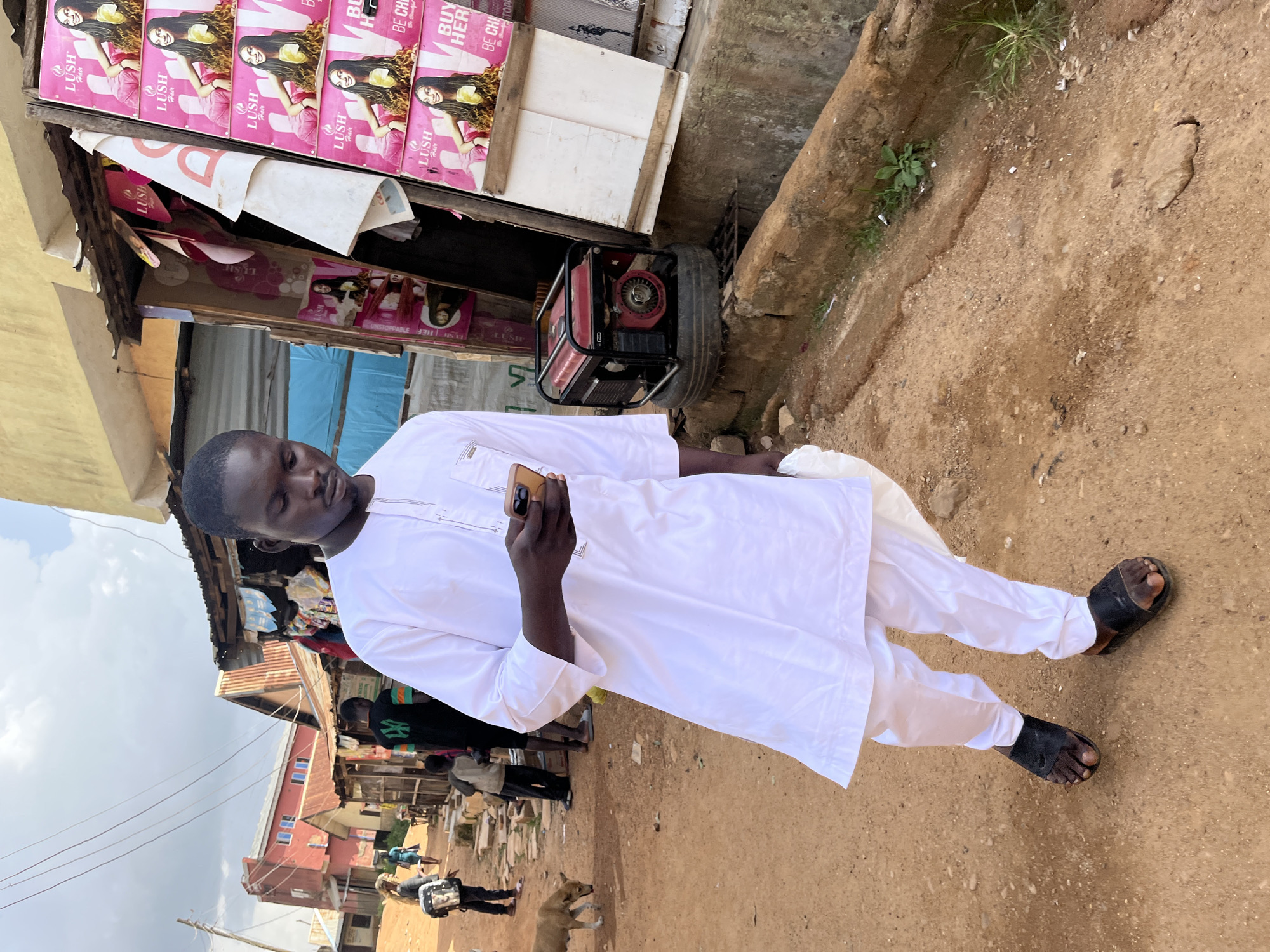A young man dressed in a white kaftan and trousers walks while looking at his phone in a dusty street in Jos, Nigeria. He wears black sandals. Behind him, other people and a red building are visible. To his right, a shop displays pink posters featuring images of women and text that appears to be advertising hair products. A red generator sits on a tire next to the shop. The ground is a mix of sand and dirt, with some rocks and sparse vegetation. The sky is cloudy.