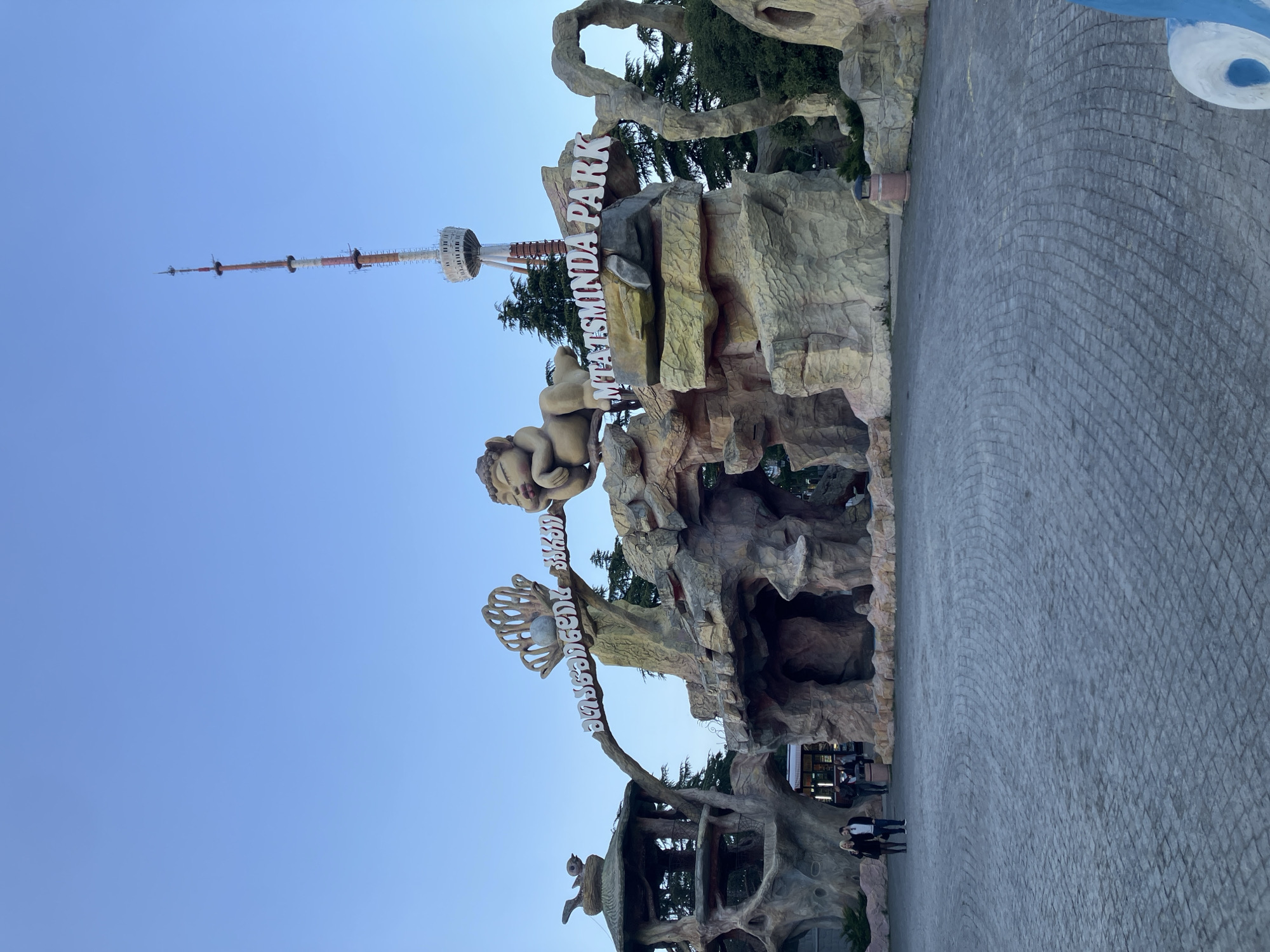The image captures the entrance to Mtatsminda Park in Tbilisi, Georgia, under a clear, bright blue sky. A large, ornate sculptural structure, designed to resemble natural rock formations and tree branches, forms the park's gateway. Prominently displayed on this structure are the words "MTATSMINDA PARK" in white capital letters, along with the same name in Georgian script ("მთაწმინდა პარკი"). A large, whimsical statue resembling a baby or cherub is positioned on the rock-like formation above the Georgian text. Green trees are visible behind and around the entrance, suggesting a natural and recreational setting.
In the background, against the vibrant blue sky, the distinctive Tbilisi TV Tower stands tall. The tower features multiple sections and a visible viewing platform. The ground leading up to the park entrance is a wide, paved area made of grey, interlocking bricks arranged in an intricate, curving pattern. A few people are seen walking on this paved path, indicating visitor activity. The scene suggests a clear, sunny day, likely during late morning or afternoon, at a popular recreational destination.
