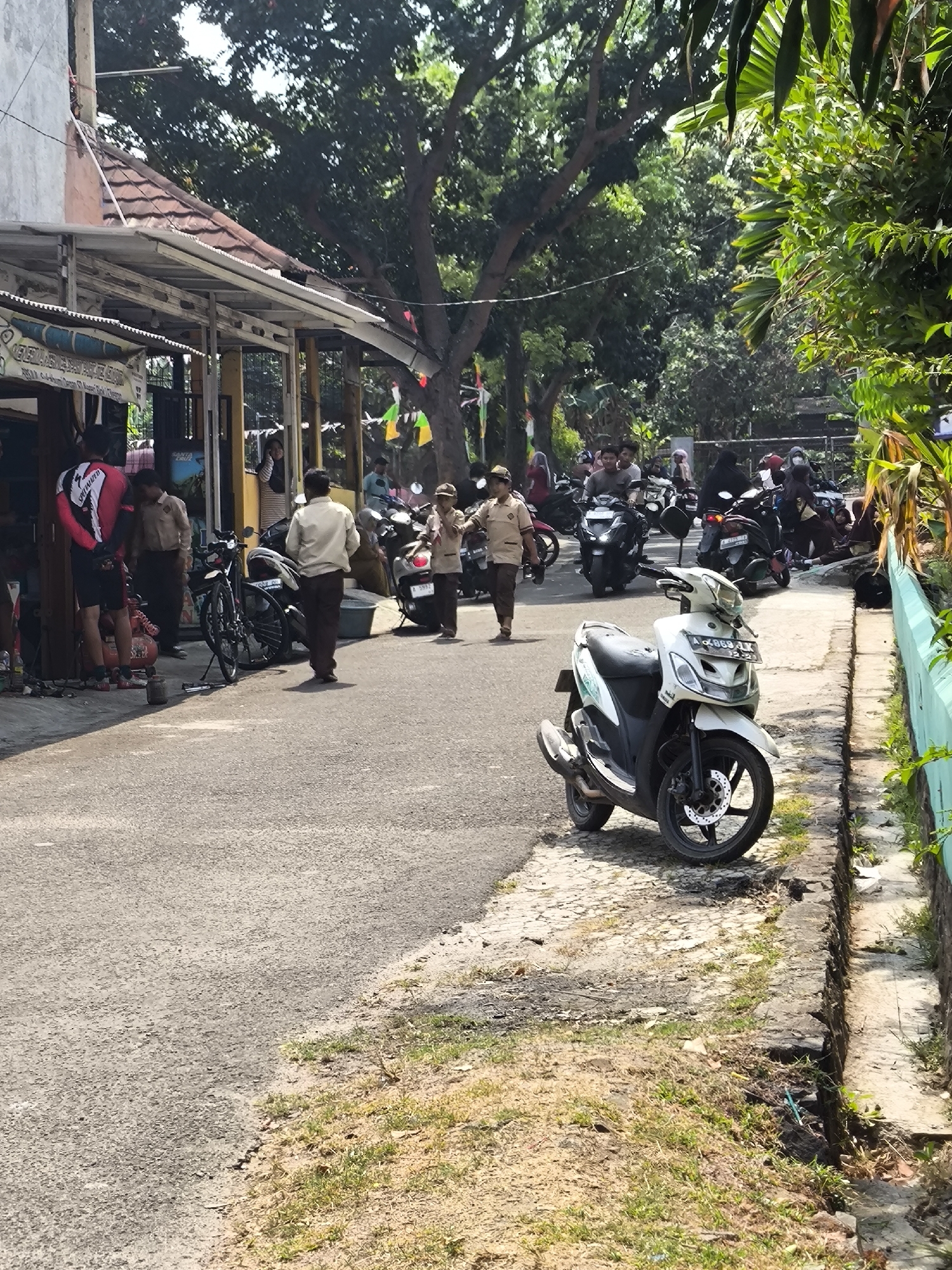 This is an outdoor street scene in Cilegon, Indonesia, likely in a residential or commercial area with a road and sidewalk. In the foreground, a white and green scooter is parked on the side of the road. The road is asphalt and appears to be a public thoroughfare. To the left of the road is a building with a covered awning, possibly a shop or workshop, with several motorcycles parked in front. A person wearing a red and black cycling outfit is standing near the building. Further down the street, several individuals are walking, some in what appear to be school uniforms. A variety of motorcycles are parked and in motion along the street. Lush green trees and foliage are visible, suggesting a tropical or sub-tropical environment. The overall atmosphere appears to be a typical day with people going about their activities.