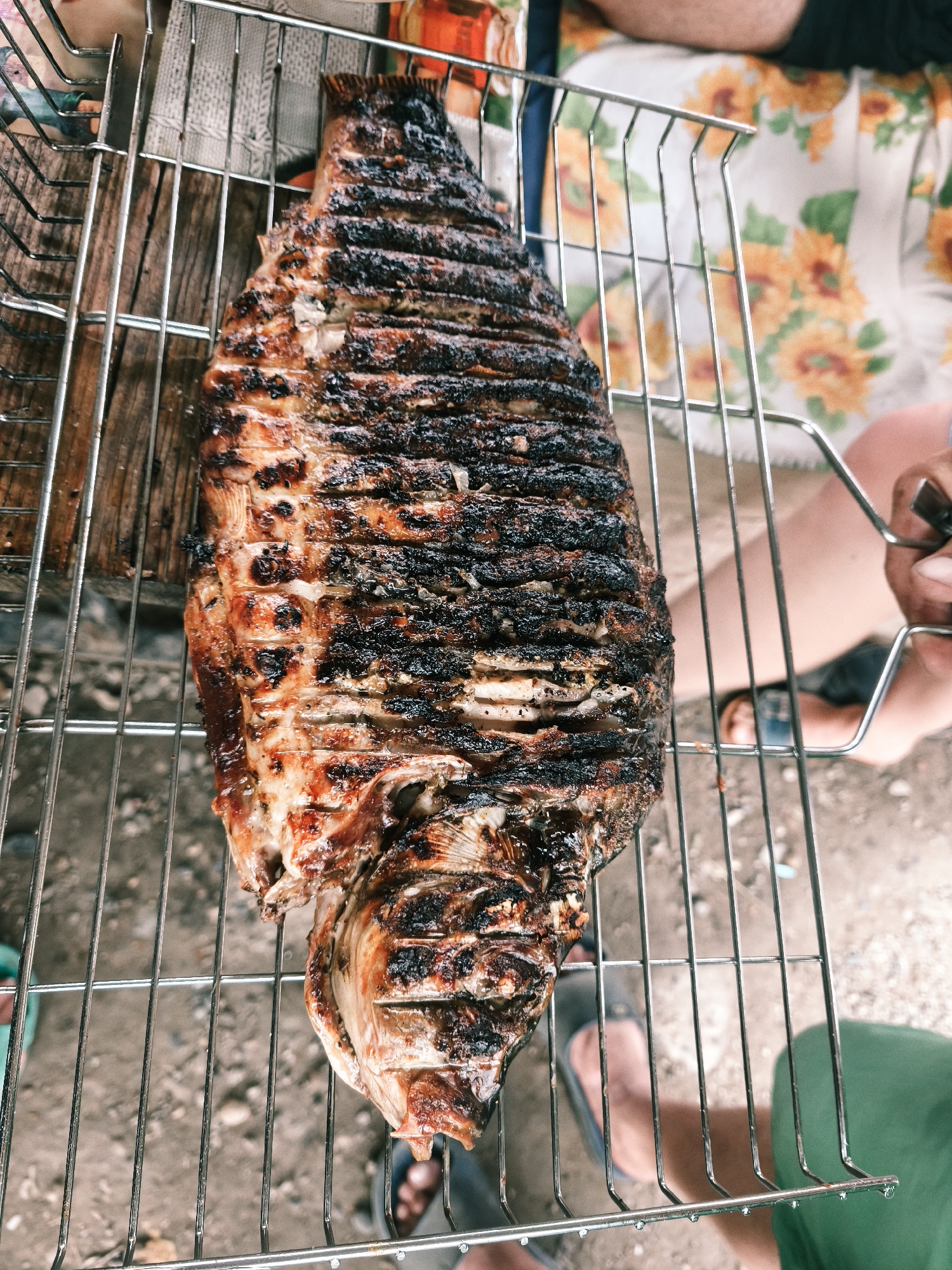 An overhead, close-up shot captures a whole, grilled fish resting on a metal barbecue grate. The fish, with its skin charred and flaky, appears to be positioned for cooking or serving. The grate itself is made of thin, parallel metal bars, supporting the fish evenly. Below the grate, a rough, textured surface, possibly dirt or gravel, is visible. In the background, partially obscured, are elements suggesting an outdoor setting: a wooden surface, the legs and feet of individuals, and a piece of fabric with a floral pattern. The lighting is natural, indicating daylight. The image is devoid of any specific landmarks or indicators that would place the scene in Nevinnomyssk, Russia, or any other particular city.