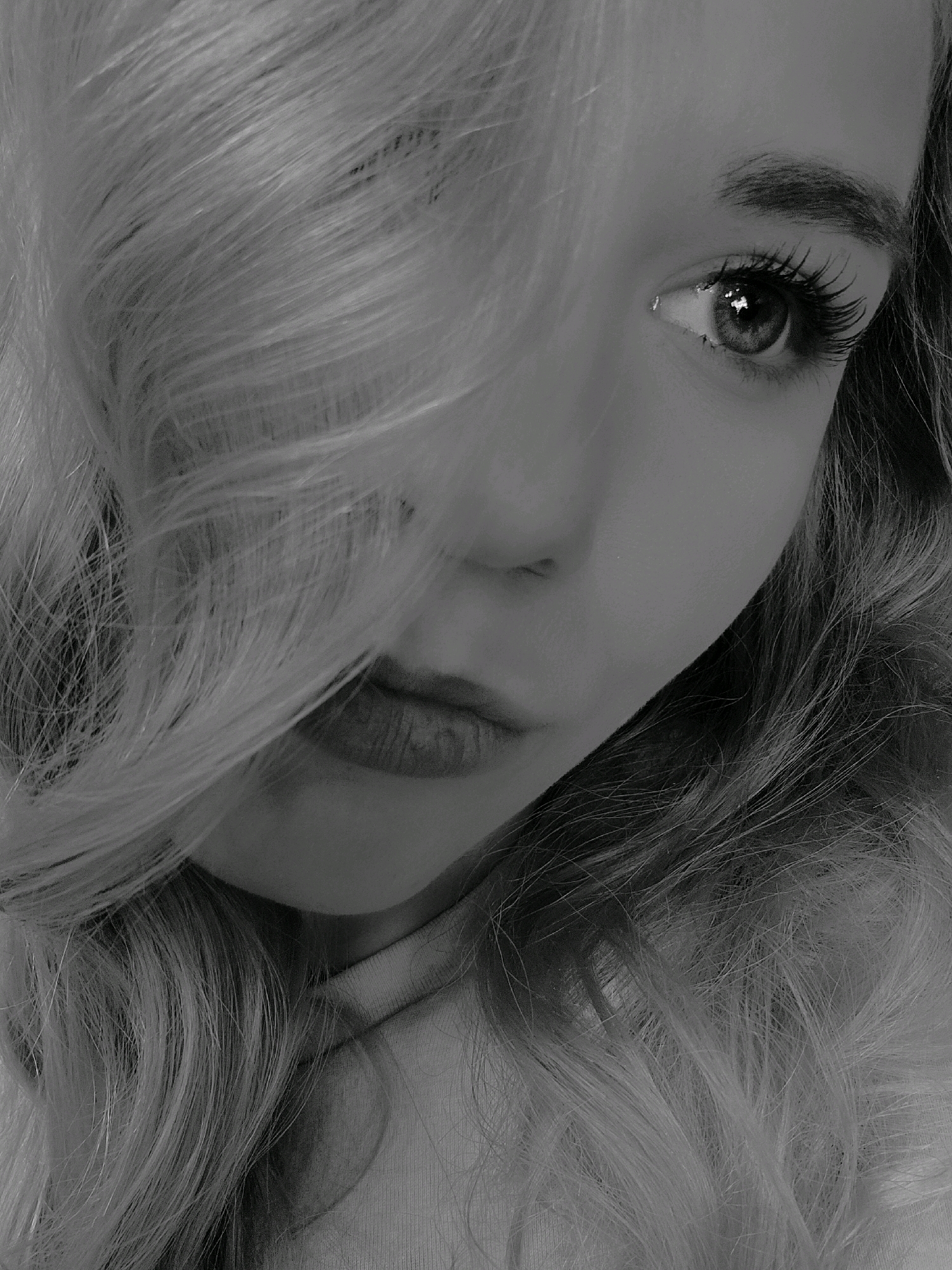 The image is a close-up, black and white photograph of a young woman's face, partially obscured by her blonde hair. Her left eye is clearly visible, with long eyelashes and a reflection in the pupil. Her lips are slightly parted and appear to be wearing lipstick. The background is indistinct and does not provide any specific environmental clues. There is no information in the image that allows for the identification of a location in Pazardzhik, Bulgaria.