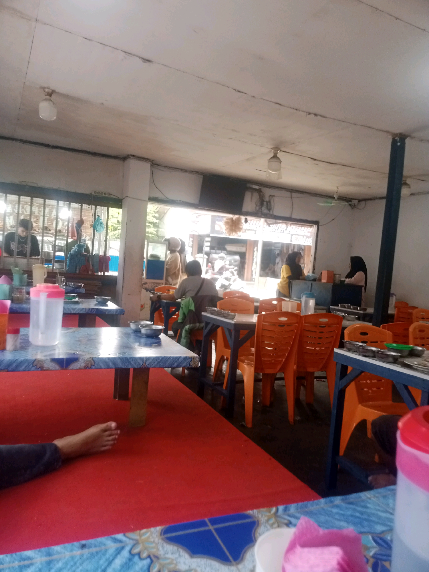 The image depicts the interior of a humble eatery, possibly a warung, in Pekanbaru, Indonesia. The scene is dominated by blue and orange plastic tables and chairs, arranged in a casual dining setting. A red carpet covers a portion of the floor. The ceiling is constructed of what appears to be a rough, unfinished material, with exposed wiring and a couple of light fixtures. In the background, a counter area is visible, where two women, one wearing a yellow shirt and the other a dark hijab, are attending to customers or preparing food. Through an open doorway or large window, a glimpse of the outside world is visible, suggesting it's daytime. A young man is seen standing behind a counter in the left background, and another person, possibly a customer, is seated at a table in the middle ground, with their back to the camera. A bare foot is visible on the red carpet in the foreground, suggesting someone is relaxing or perhaps working in the establishment. The overall impression is of a bustling, no-frills establishment catering to local patrons.