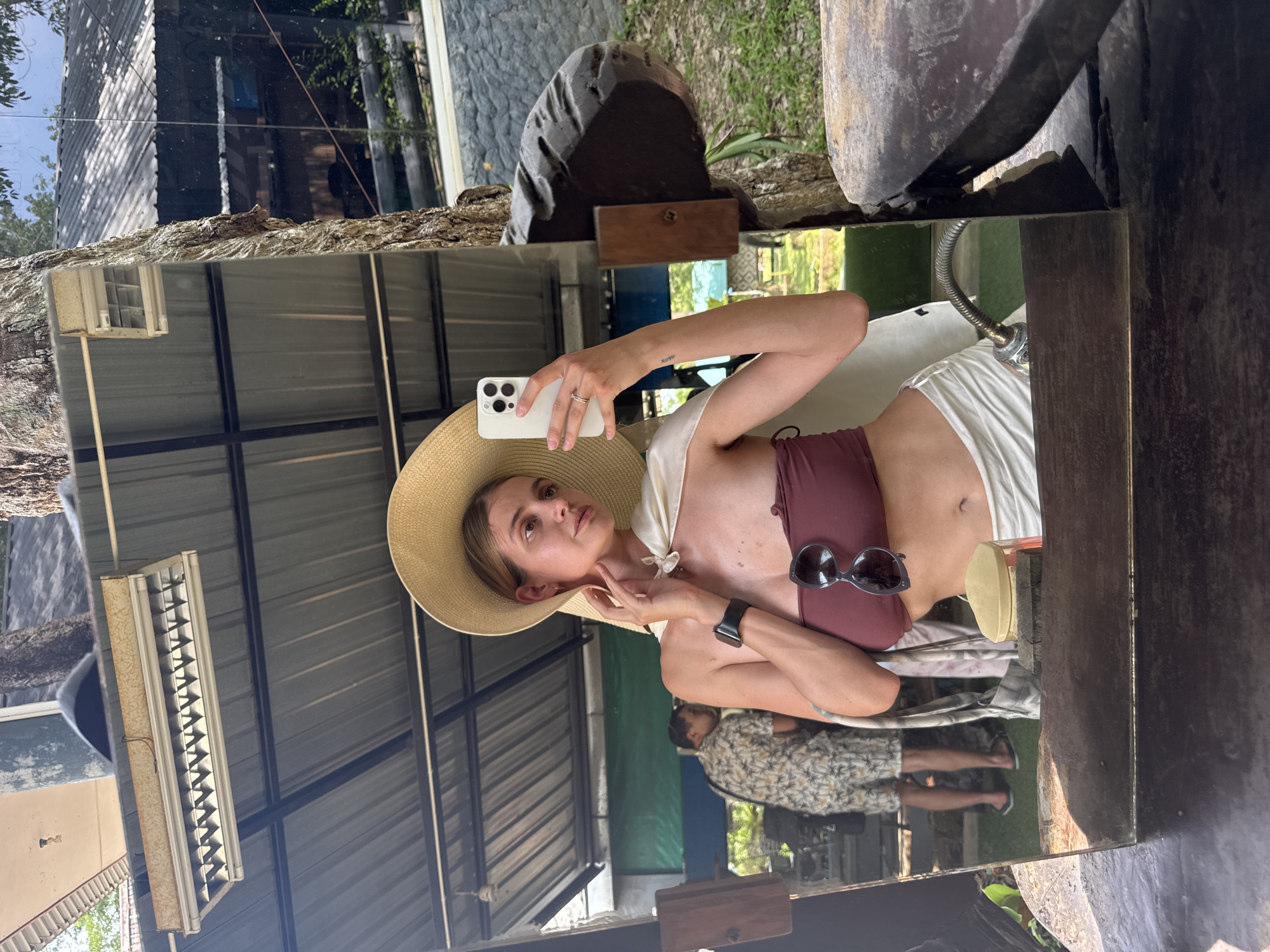 A woman wearing a large straw hat, a maroon bikini top, and a white cover-up is taking a selfie in a mirror outdoors. Sunglasses rest on her bikini top, and she wears a watch on her left wrist. She holds a white phone with her right hand. The mirror reflects the underside of a metal awning, a man sitting at a table, and outdoor greenery. The location is Ban Khlong Khian บ้านคลองเคียน, Thailand.