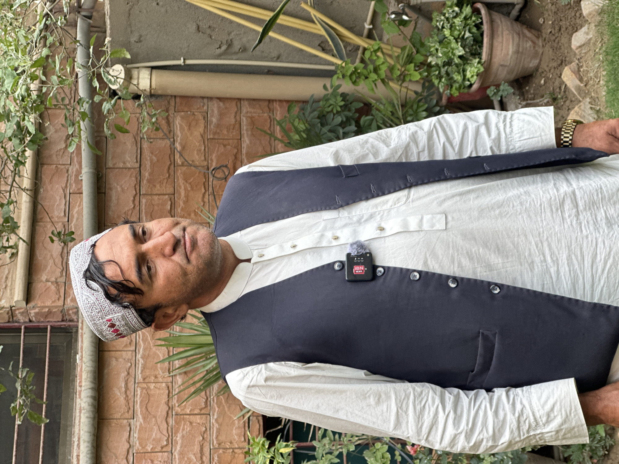 A man stands in what appears to be an outdoor area in Quetta City Tehsil, Pakistan. He wears a white shalwar kameez with a navy waistcoat and a traditional Pakistani cap. A microphone with an "IBN News" logo is clipped to his waistcoat. He is wearing a gold wristwatch on his left wrist. The background shows a brick wall, various plants, and a possible building structure. The area seems like a garden or courtyard.