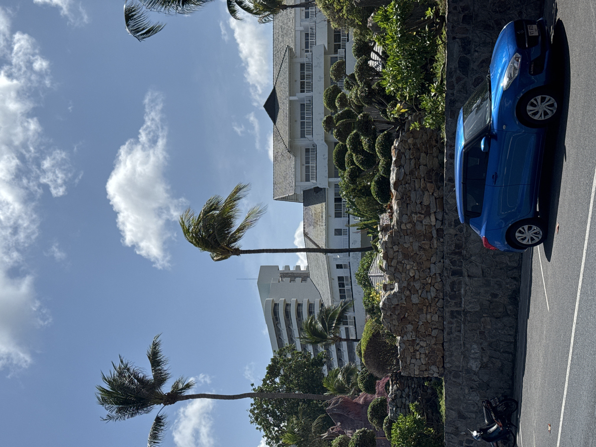 The image shows a bright, sunny day in Karon, Thailand. A blue car is parked in the foreground on an asphalt road with white parking lines. To the left of the car, a black motorcycle is parked. In the background, there is a stone wall, lush green landscaping with manicured bushes, and two white buildings. One building is a multi-story hotel with balconies, and the other appears to be a resort or another hotel with a tiled roof. Several palm trees are visible, including two tall ones framing the buildings. The sky is blue with scattered white clouds.