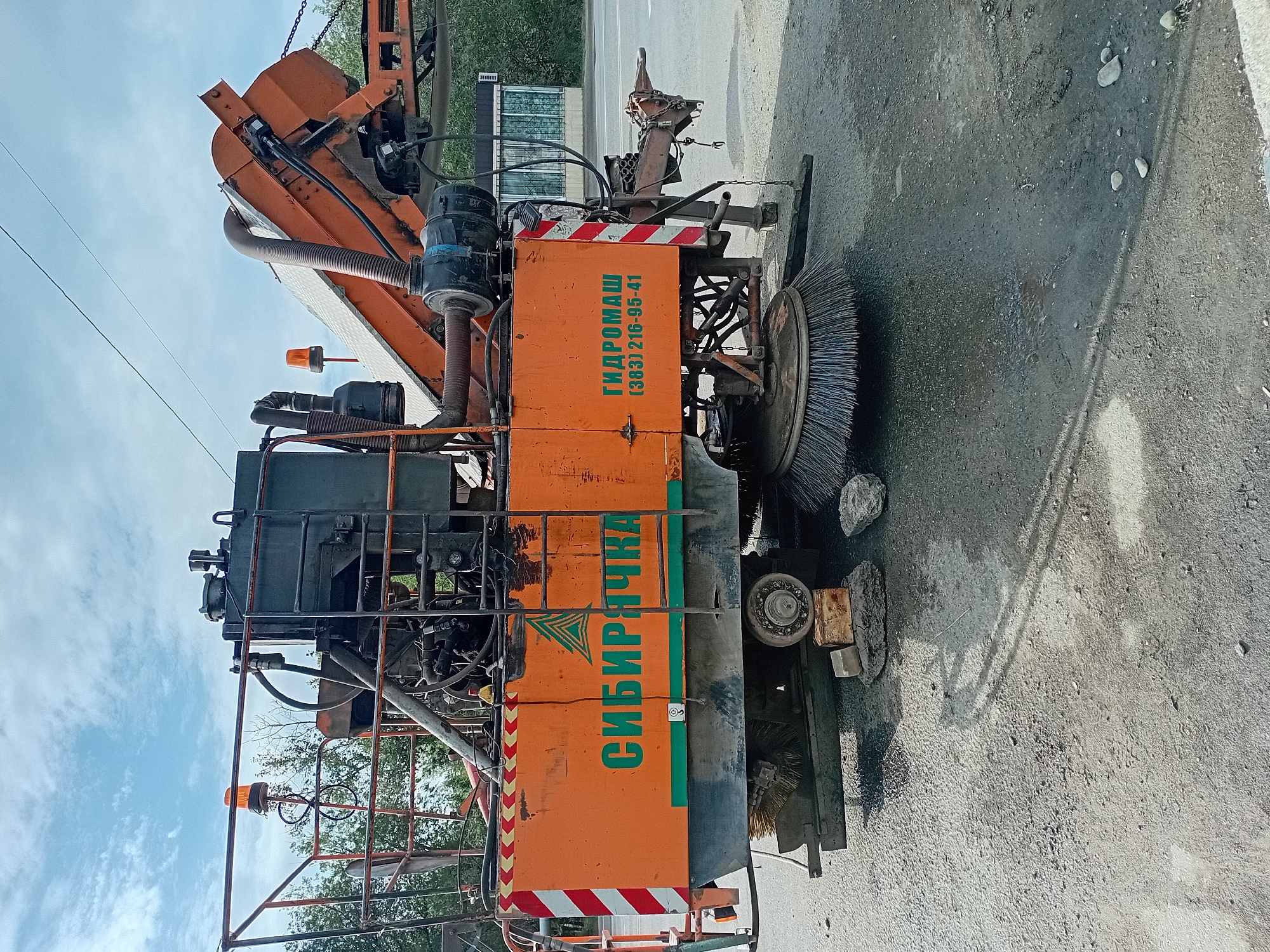 The image shows a street sweeper vehicle parked on a paved road, likely in the city of Semey, Kazakhstan. The vehicle is predominantly orange and dark gray, with visible mechanical components, a large rotating brush, and an orange warning light. Text on the side of the vehicle reads "СИБИРЯЧКА" and "ГИДРОМАШ (383) 216-95-41". The background includes trees, a cloudy sky, and a small building. The road surface appears worn and dusty, with some debris and rocks present. The vehicle appears to be stationary.