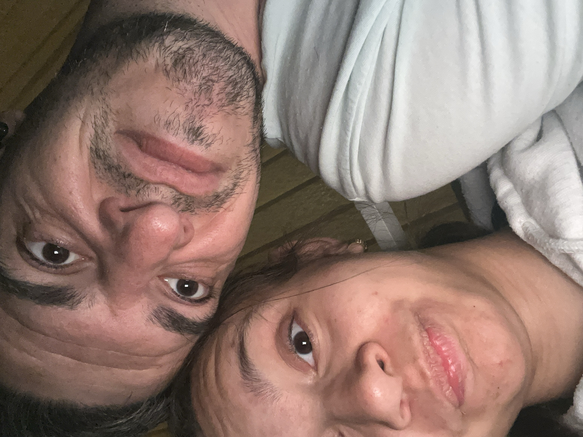 The image is a close-up selfie featuring two individuals, a man and a woman. The man's face is positioned above and to the right of the woman's face, with his head tilted. Both individuals are looking towards the camera. The man has dark hair, a beard, and appears to be in his late 30s or 40s. The woman has dark hair and appears to be in her 20s or 30s. They are both wearing casual clothing. The background appears to be indoors, with a wooden-paneled wall visible behind them. The image quality is slightly grainy, suggesting it was taken in low light conditions. There is no specific information in the image that indicates the location is the city of Sofia, Bulgaria.