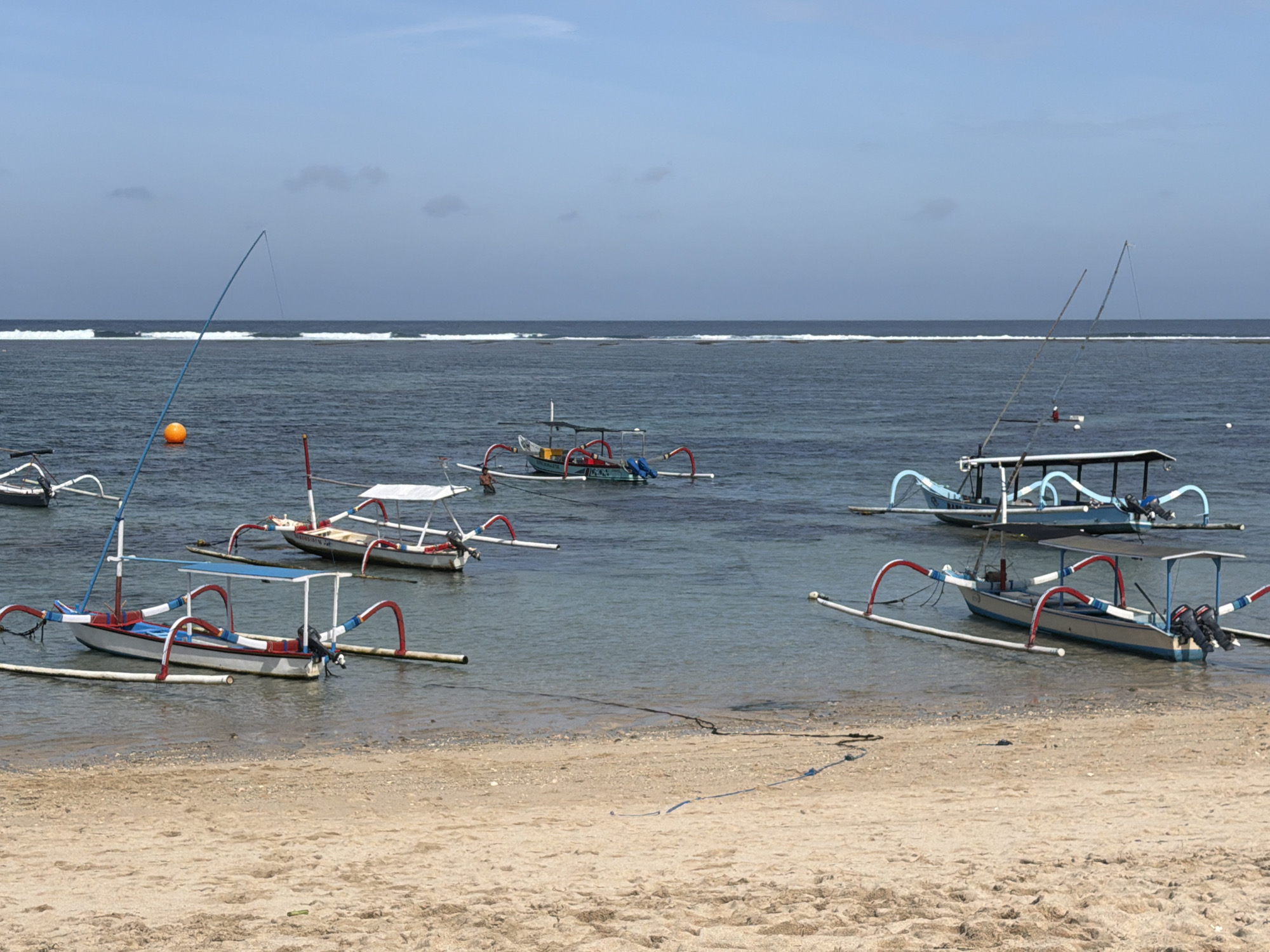 A beach scene in Kuta Selatan, Indonesia, depicts several outrigger canoes floating on the calm, blue water. The sand in the foreground is light brown and appears smooth, with some small pebbles scattered across it. The water is shallow near the shore, becoming deeper as it extends towards the horizon, where a line of white waves breaks against a reef.
In the foreground, several outrigger canoes are moored. The closest canoe, on the left, is white with red and blue accents on its outriggers and is equipped with a blue canopy. Another canoe, further back and slightly to the right, also features red and white outrigger supports and a white canopy. A third canoe, positioned in the middle ground to the right, is blue with white trim and has two outboard motors attached to its stern. Several other canoes, less distinct due to distance, are visible further out on the water.
In the water, to the left of the nearest canoe, a bright orange buoy is visible. A long, thin fishing rod, its tip bent towards the water, extends from the shore towards the buoy and the fishing grounds. The sky is a pale blue with a few scattered, wispy clouds. The overall impression is a serene coastal setting with traditional fishing vessels.