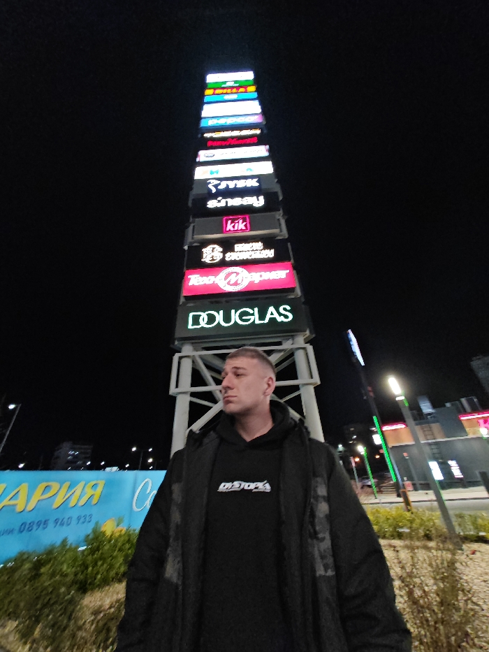 The image displays an outdoor nighttime scene at Retail Park Shumen, located at бул. "Симеон Велики" 116, 9700 Shumen, Bulgaria. A male subject is positioned in the foreground, slightly right of center, looking towards his left with a neutral expression. He has a short, faded haircut and is dressed in a dark hooded sweatshirt featuring white text "DYSTOPIA," worn under a dark, possibly quilted, jacket.
Behind the subject, a prominent, tall, illuminated sign tower rises against the dark sky. This tower, constructed with a light-colored metal framework, displays multiple stacked, rectangular signs for various businesses. Visible logos and names, from bottom to top, include DOUGLAS, TeoMаркет, ЕДОН СГЛОБЯВАНЕ (EDON SGLOBYAVANE), kik, sinsey, JYSK, Pepco, Mr. Bricolage, and BILLA. To the left of the male subject, a partial blue billboard is visible at ground level, displaying Cyrillic text and a Bulgarian phone number (0895 940 933). Low-lying green foliage partially obscures the billboard. In the background to the right, additional commercial buildings with illuminated facades and tall, vertical streetlights emitting white and green light are discernible. The environment is illuminated entirely by artificial light sources from the commercial signs and streetlights, set against a dark night sky.