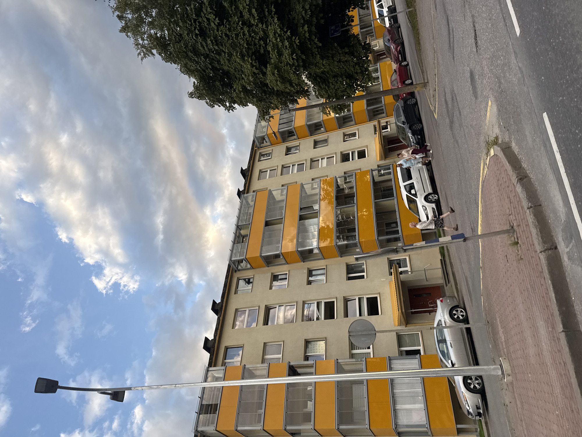 The image depicts a street scene in Tallinn, Estonia, likely during the late afternoon or early evening, given the lighting. A multi-story apartment building with bright yellow balconies dominates the left side of the frame. The building appears to be made of concrete with yellow accents. Several cars are parked on the street and a few pedestrians are visible.
On the left side of the image, a tall, dark streetlamp stands prominently. In front of the building, a silver sedan is parked. Further down the street, a white van is parked, with three people standing near it. One person, a male in a light blue shirt and white shorts, is interacting with a female in a beige top and dark pants. Another person, a female wearing a white jacket and dark skirt, is walking across the street.
To the right of the building, a large, dark green tree partially obstructs the view. A few other cars, including a dark SUV, a red sedan, and another red car, are parked along the street. A white van is also visible in the distance. The street is paved with asphalt, and a pedestrian walkway made of red bricks is present. The sky is partly cloudy, with a mix of blue sky and gray clouds. The overall impression is of a residential area in a European city.
