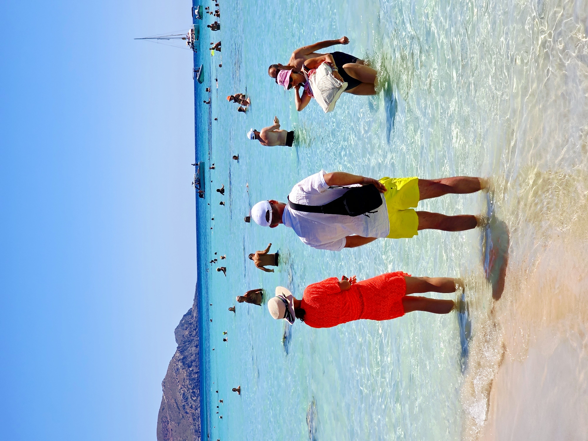 A bright, sunny day at a crowded Greek beach is depicted. In the foreground, a couple stands in shallow, crystal-clear turquoise water. The woman wears a vibrant orange dress and a straw hat, while the man is dressed in a white shirt and bright yellow shorts, with a black bag slung across his chest. Behind them, other beachgoers are visible, some swimming, some standing in the water, and others with boats in the background. The water is incredibly clear, allowing the sandy bottom to be seen, and gentle waves lap at the shore. The backdrop features a mountainous landscape under a clear blue sky.