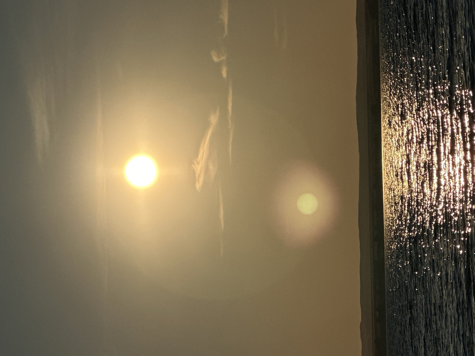 The image depicts a serene sunset over a body of water, likely a reservoir, as the sun is low on the horizon, casting a warm golden light. The sky is a hazy yellow-orange, with a few wispy clouds. There are two suns visible, likely an optical illusion caused by atmospheric conditions. The larger sun is in the upper part of the frame, while a smaller, fainter sun appears below it. The water in the foreground is dark blue, with the sunlight creating a shimmering reflection on its surface. In the background, a dark, silhouetted landmass, possibly hills or mountains, forms the horizon. This scene is consistent with the natural beauty of the area surrounding Energetik, Russia, a city known for its proximity to the Sayano-Shushenskaya Dam and its associated reservoir, which would provide such a large expanse of water. The image captures a moment of natural tranquility, with no people or man-made structures prominently featured.