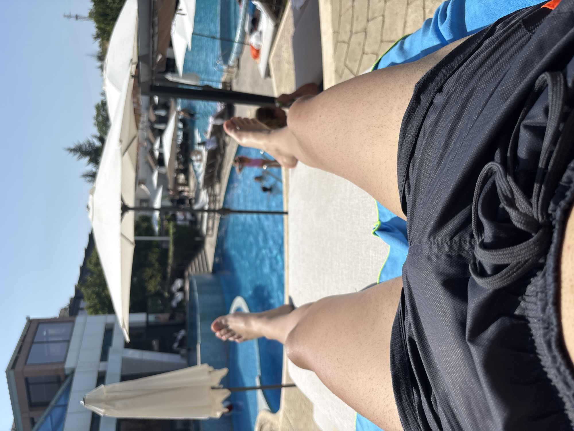 The image shows a first-person perspective of a person lounging on a recliner by a swimming pool. The person is wearing dark-colored swim shorts. Their legs and feet are visible in the foreground, with the right leg bent at the knee and the left leg extended. The person's feet are bare.
In the background, a bright blue swimming pool is visible, with several people in and around it. Some individuals are swimming, while others are sitting or standing near the water. Several white umbrellas are set up around the pool area, providing shade. A modern building with large windows is situated behind the pool, and lush green trees are visible further in the background. The sky is clear and blue, indicating a sunny day. The location appears to be an outdoor resort or hotel pool area, possibly in Polenitsa, Bulgaria, given the provided context. The overall scene depicts a relaxing atmosphere.