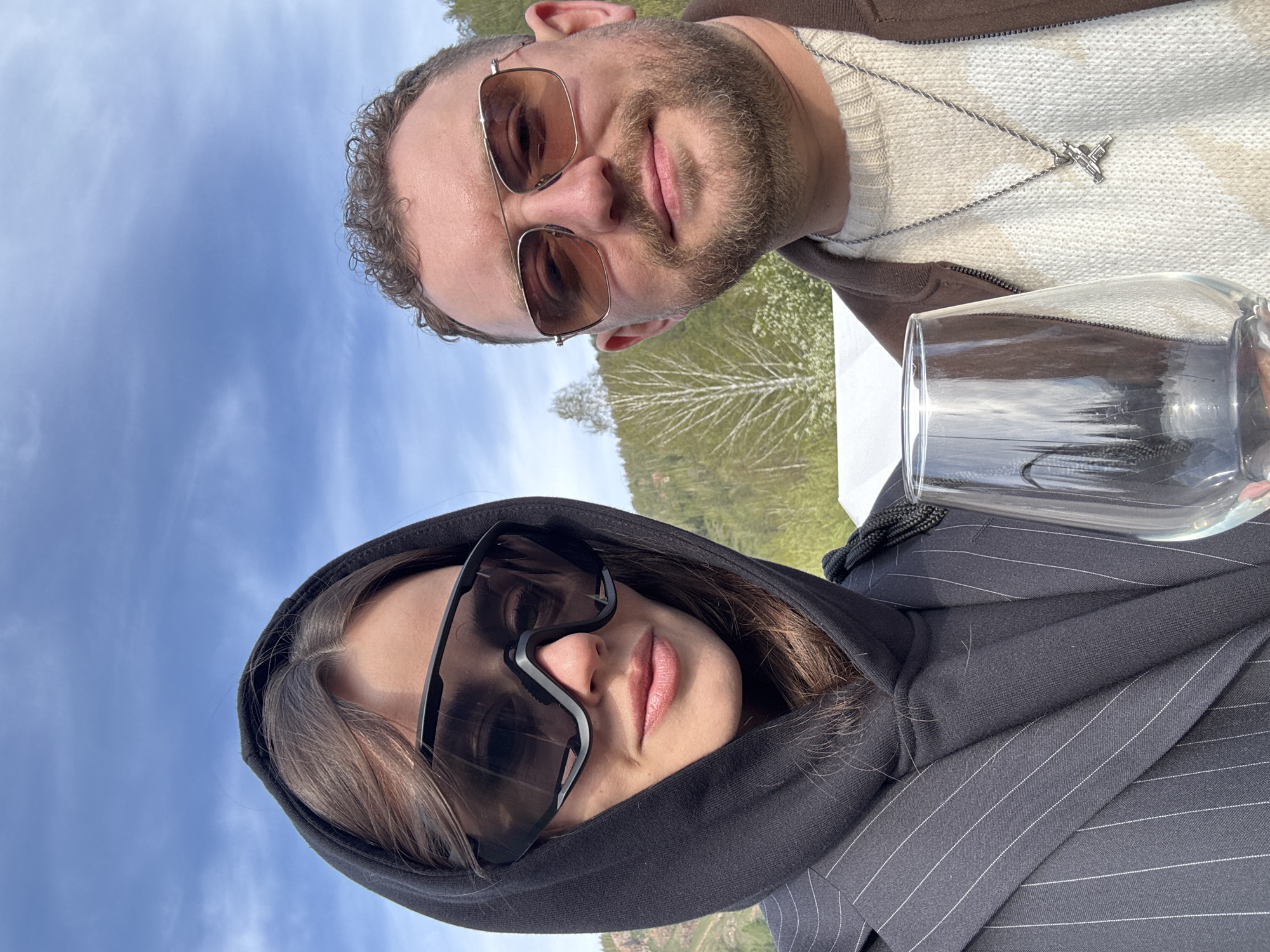 Two individuals are pictured in what appears to be an outdoor setting. The man on the top is wearing sunglasses, a brown jacket over a white patterned sweater, and a silver necklace. He has short, curly hair and a beard. A glass is positioned near him. The woman on the bottom is wearing a black hooded top with white pinstripes and large sunglasses. Behind them is a view of trees and sky, which suggests an outdoor environment, possibly within the city of Krasnoyarsk, Russia.