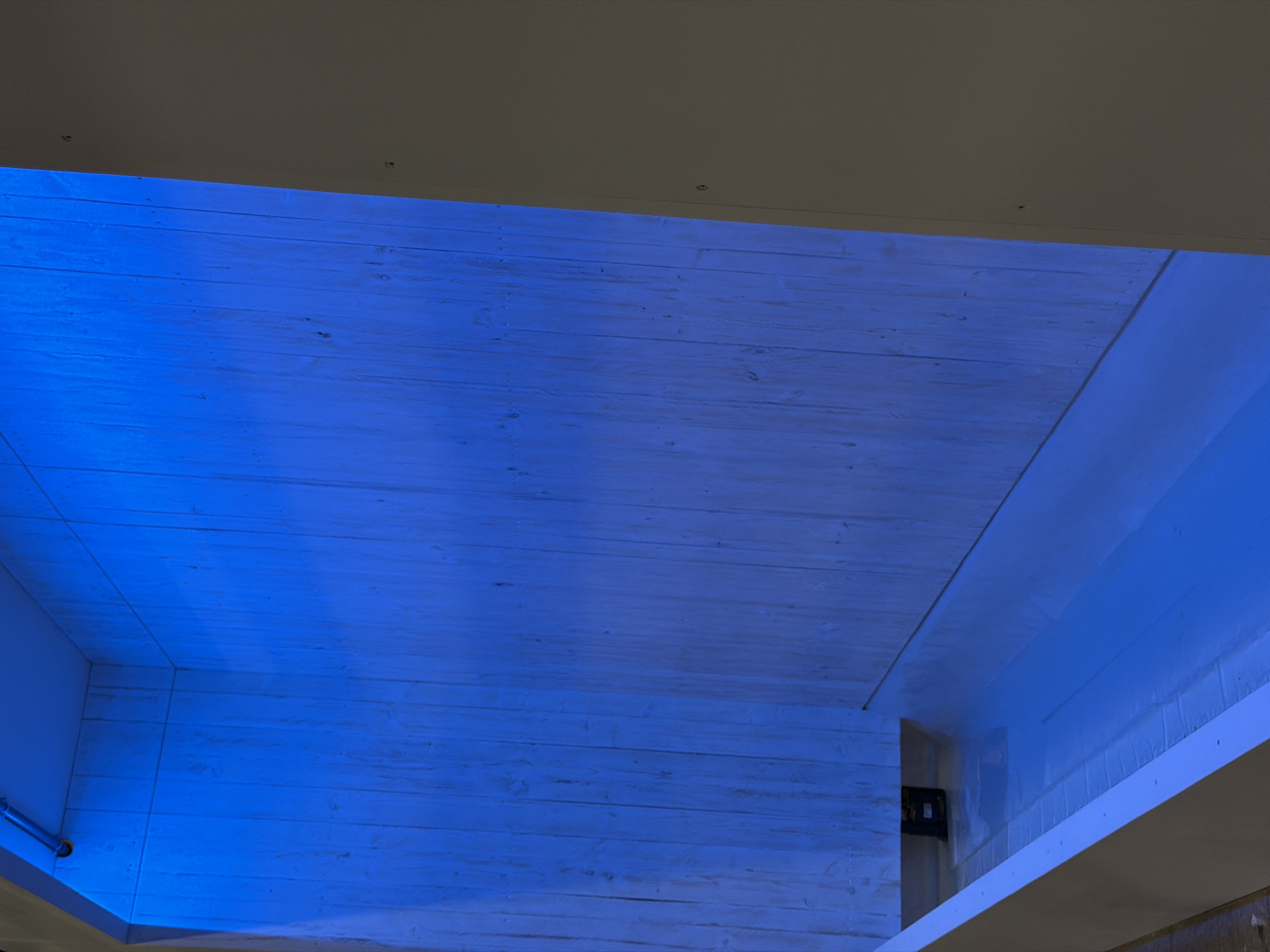 The image shows a long, narrow, rectangular channel filled with blue-tinted water, set within a shower stall. The walls of the stall are made of light-colored wooden planks, illuminated by blue LED lighting. A metal pipe is visible along the top of the stall, suggesting it might be a fixture for water or lighting. The channel is open at the top, and the water appears still. The overall scene conveys a modern and possibly therapeutic spa-like environment. No people or specific identifiable landmarks are present in the image. Without additional information or context, the exact location in Leudelange, Luxembourg, cannot be determined from the image alone.