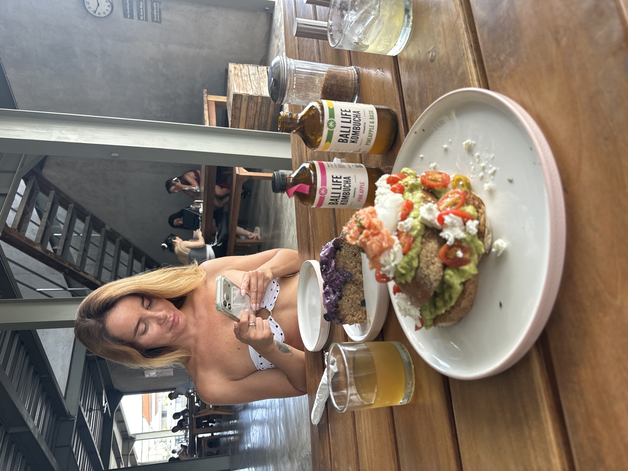 A blonde woman is seated at a wooden table in what appears to be an open-air cafe in Mengwi, Indonesia. She is looking down at her phone, which she holds in both hands. She is wearing a white and black polka dot bikini top. In front of her on the table are several items of food and drink: a slice of cake topped with blueberries, a glass of amber-colored liquid, two bottles of "Bali Life Kombucha," a sugar dispenser filled with brown sugar, and a white plate with a breakfast item topped with avocado, salmon, tomatoes, and feta cheese. In the background, other patrons are visible at tables and a staircase leads to an upper level. The cafe has a rustic, industrial aesthetic with exposed metal beams and concrete walls.