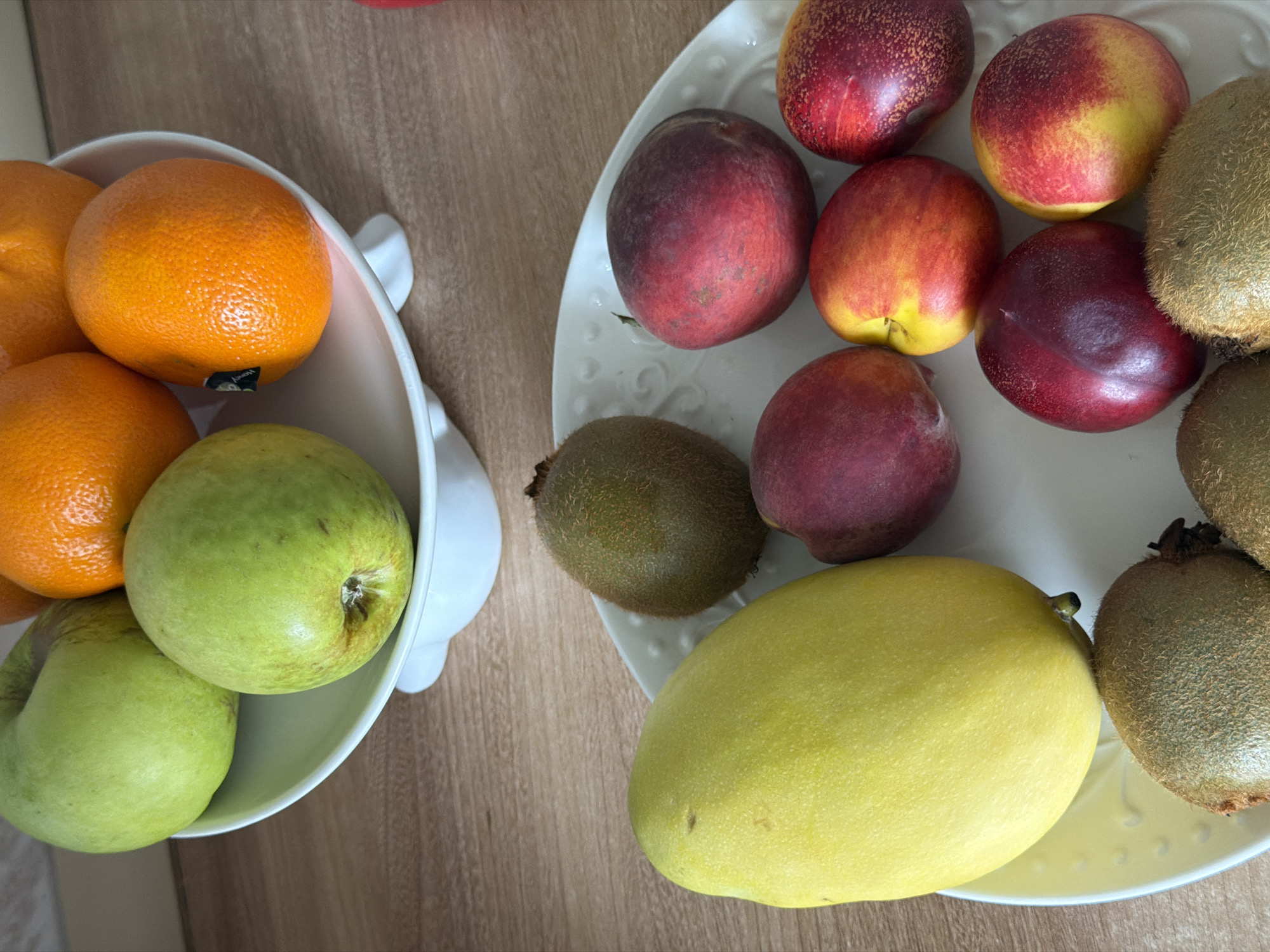 In Izhevsk, Russia, a still life arrangement of fresh fruits is presented. The scene features a white tiered fruit stand holding several green apples and oranges on the upper level. Below, on a white platter, are various fruits including a large yellow mango, several kiwis, and a collection of nectarines in shades of red, yellow, and orange. The fruits are arranged in a visually appealing manner, suggesting a domestic or culinary setting. The background consists of a wooden surface, likely a countertop or table. There are no people visible in the image.